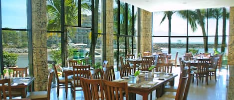 A restaurant with multiple tables and chairs, large windows offering a view of palm trees and a body of water.