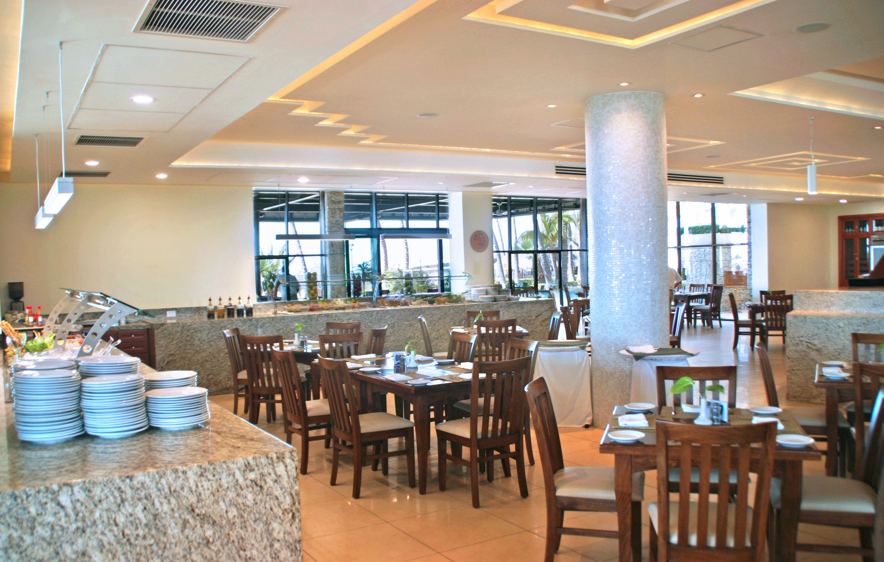 A spacious restaurant with multiple dining tables and chairs, a counter with stacked plates, and large windows providing natural light.