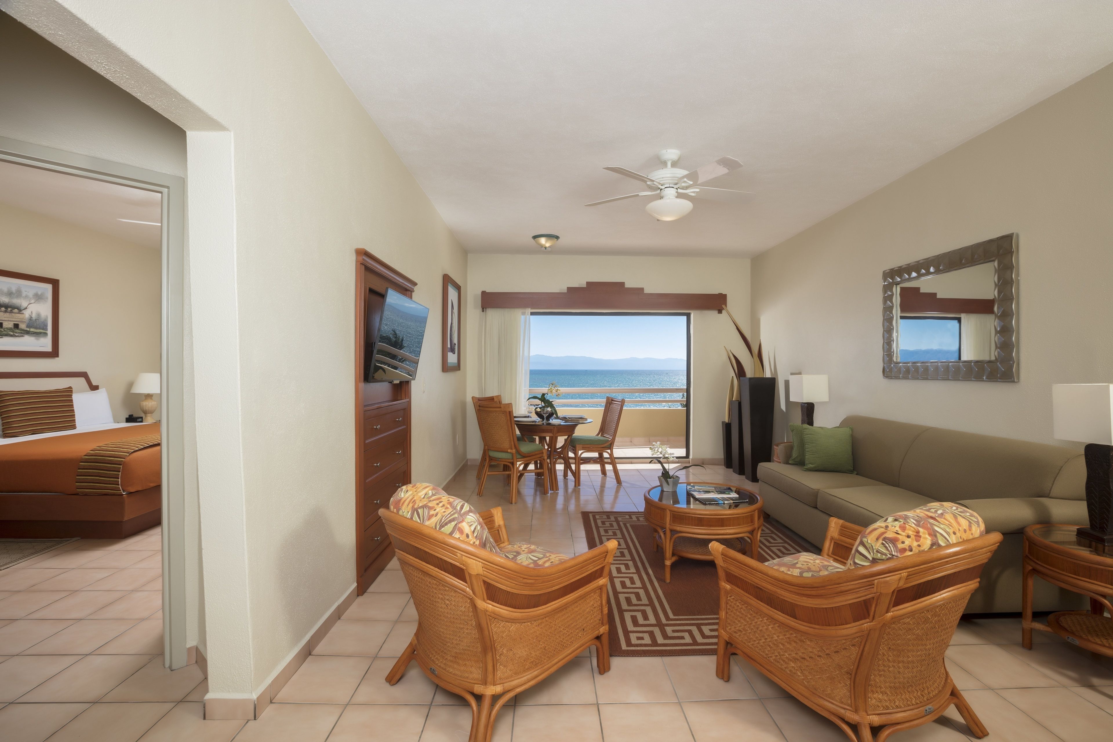 Quarto de hotel com varanda, mobiliário de vime, televisão e vista para o oceano.