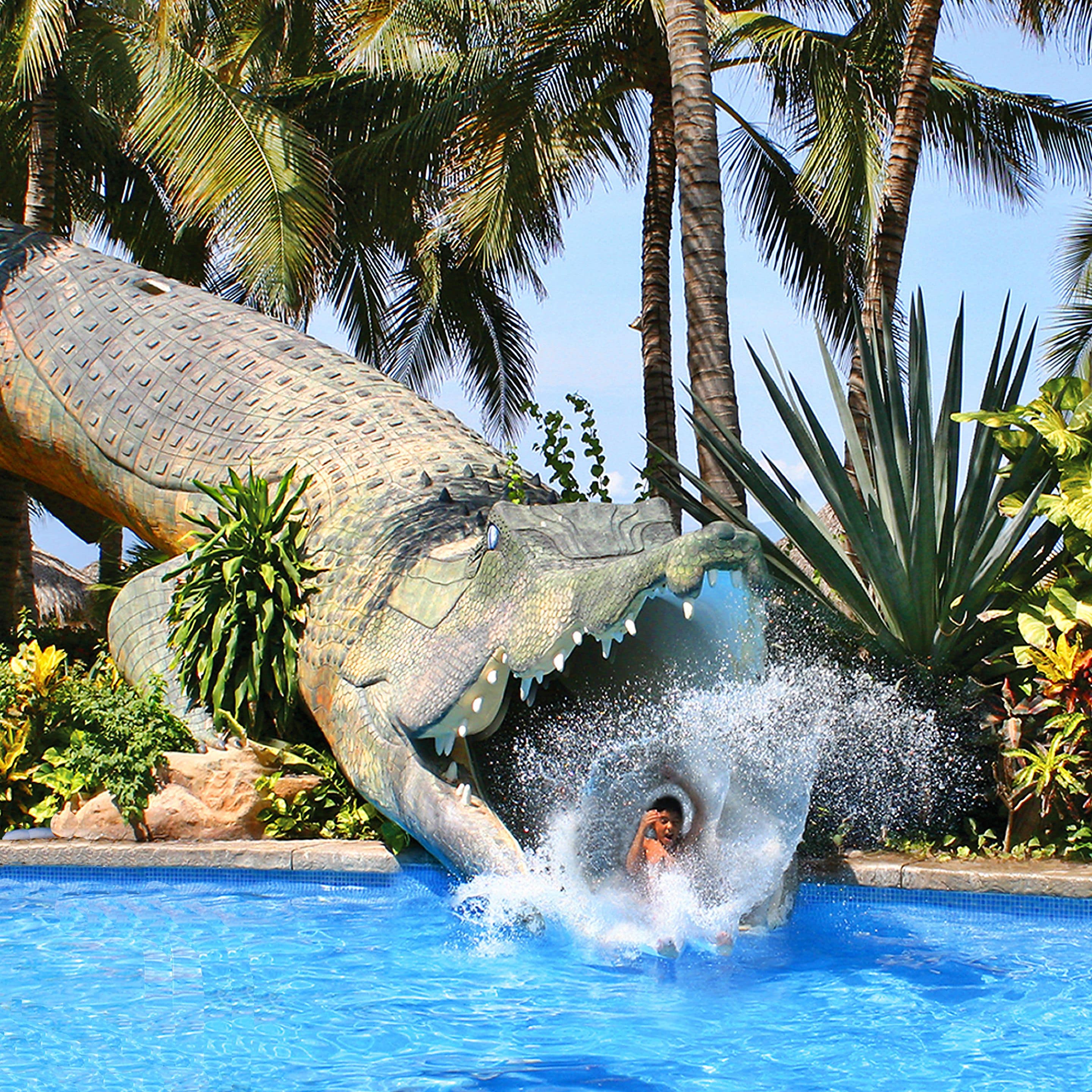 Eine große Alligatorstatue, teilweise in einem Pool versunken, mit Palmen und tropischer Vegetation im Hintergrund.
