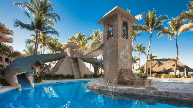 Una piscina con una escultura de dinosaurio, palmeras y una pequeña estructura tipo torre.