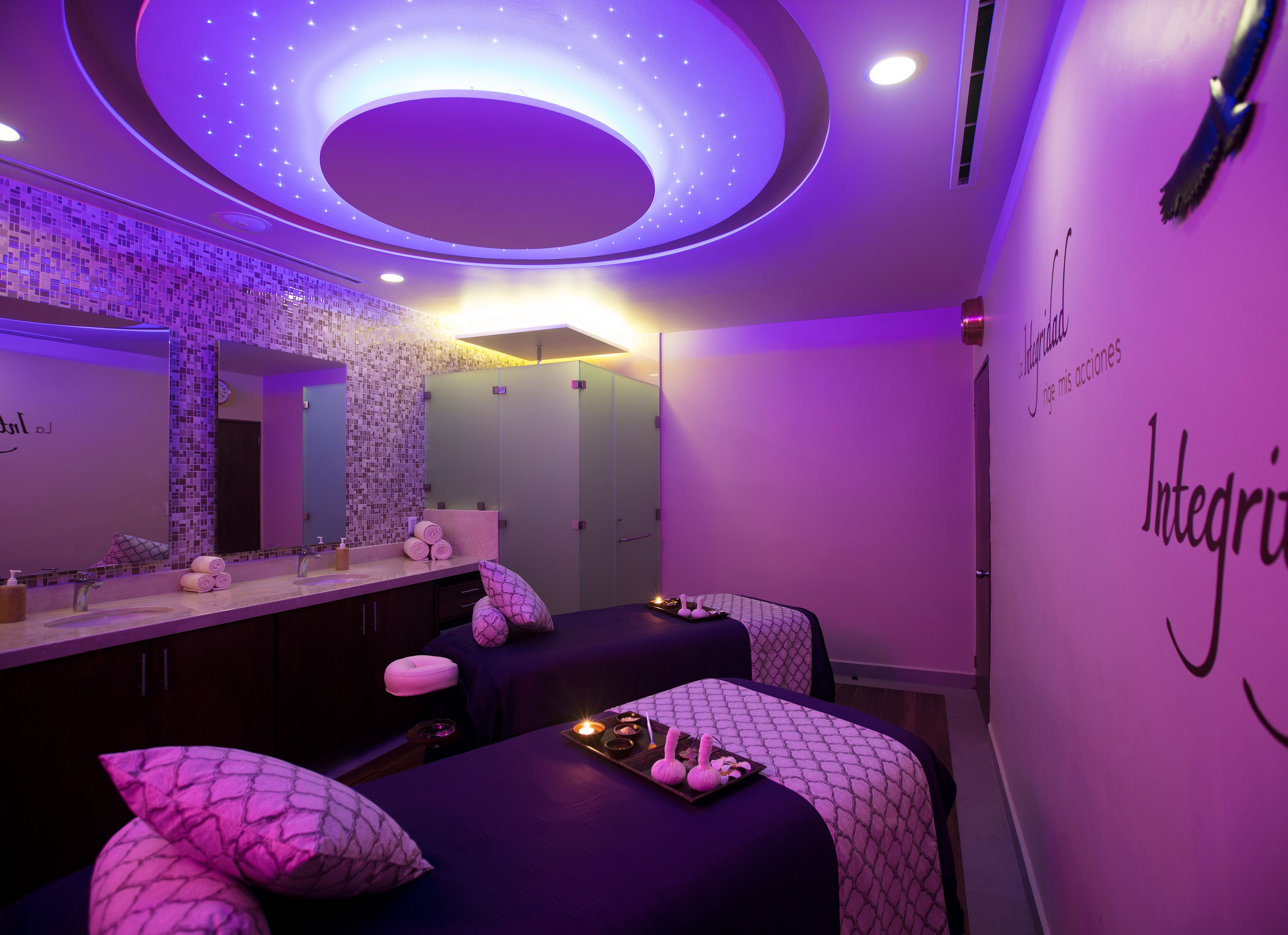 Une salle de spa équipée de deux tables de massage, d’un grand miroir et d’un plafond orné d’un luminaire circulaire.