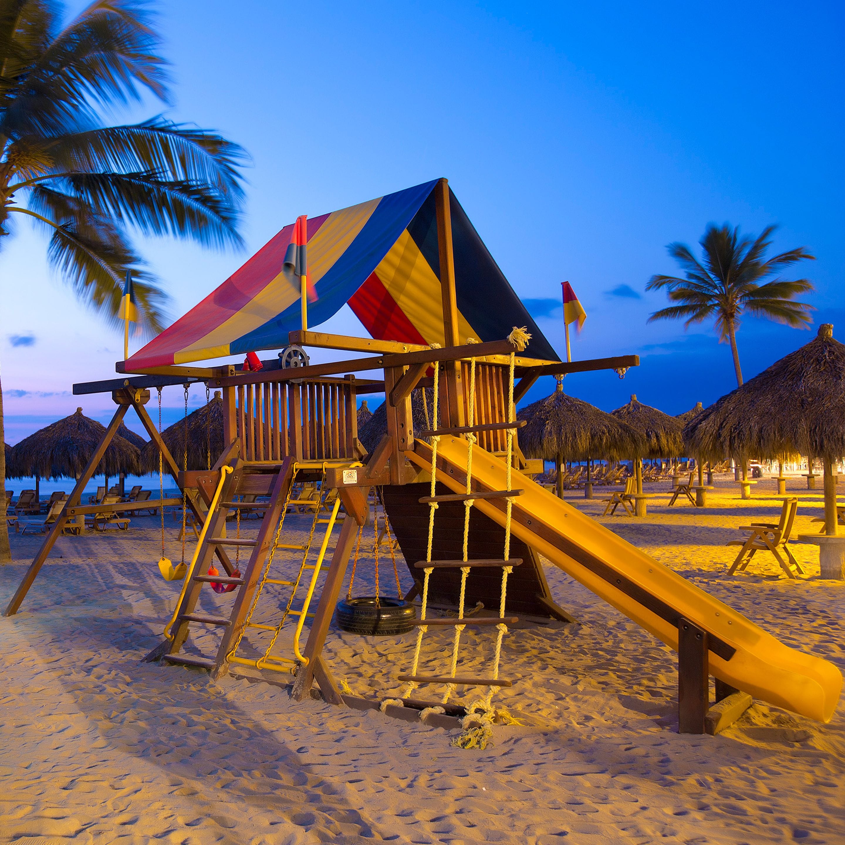 Ein Spielbereich am Strand mit Rutsche und Klettergerüst, umgeben von Palmen und Liegestühlen.