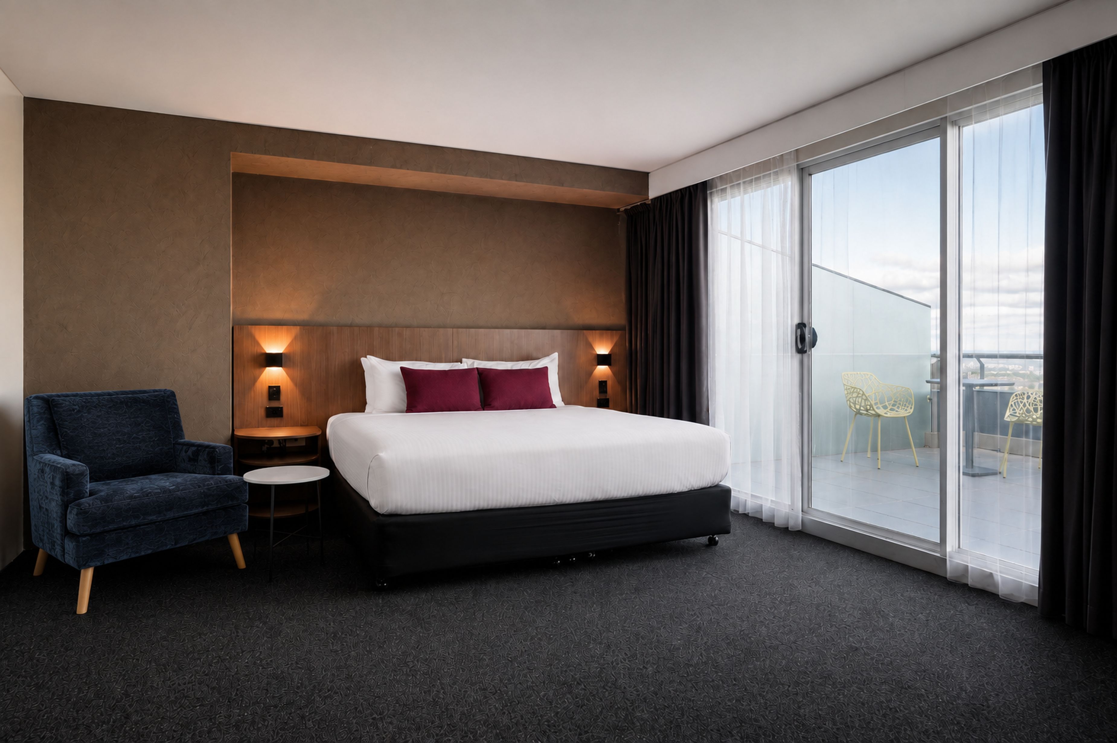 Ein Hotelzimmer mit einem großen Bett, einem Sessel, einem kleinen Tisch und Blick ins Freie durch ein großes Fenster.