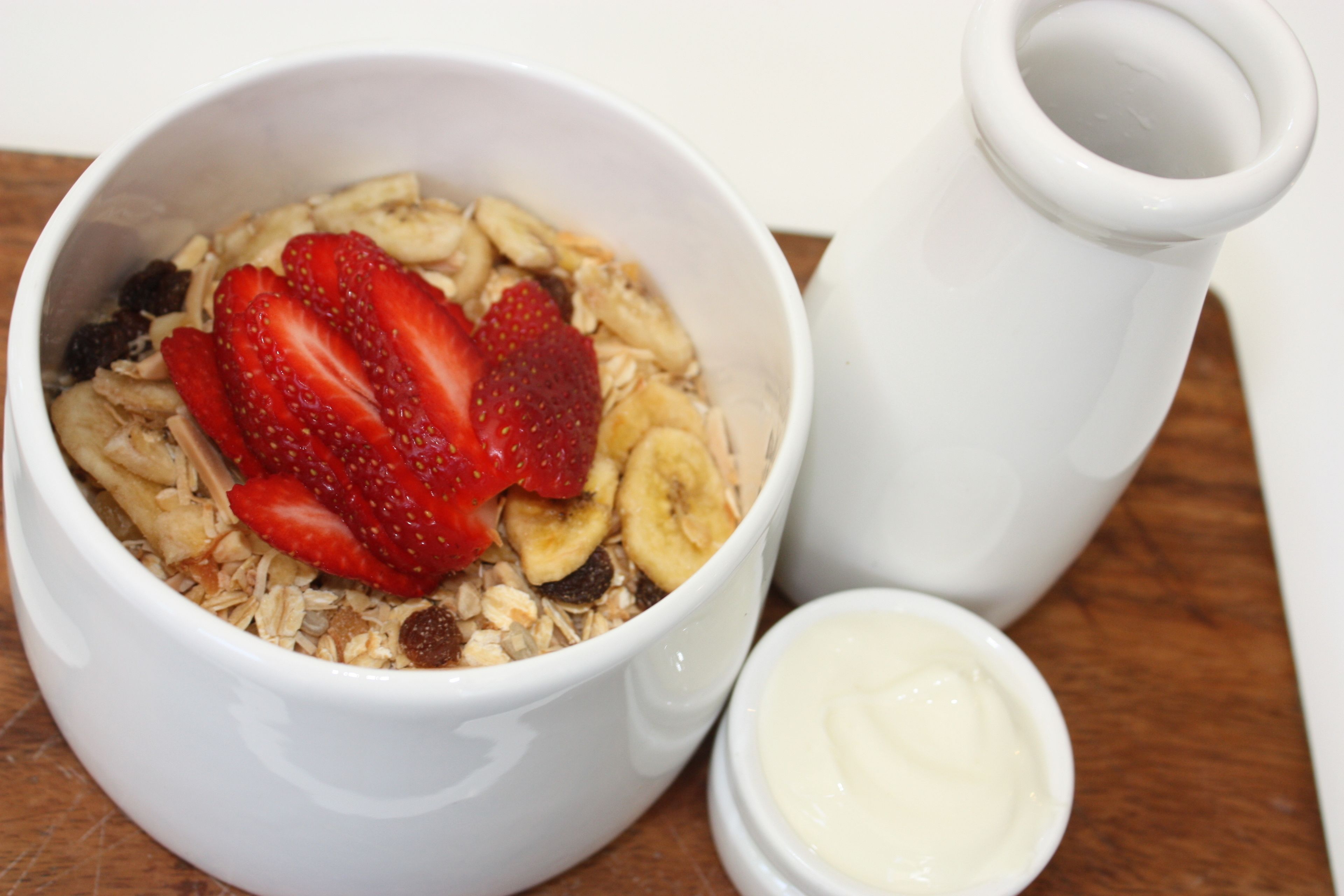 A bowl of cereal with strawberries and bananas, a small container of yogurt, and a white pitcher on a wooden surface.