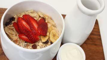 A bowl of cereal with strawberries and bananas, a small container of yogurt, and a white pitcher on a wooden surface.