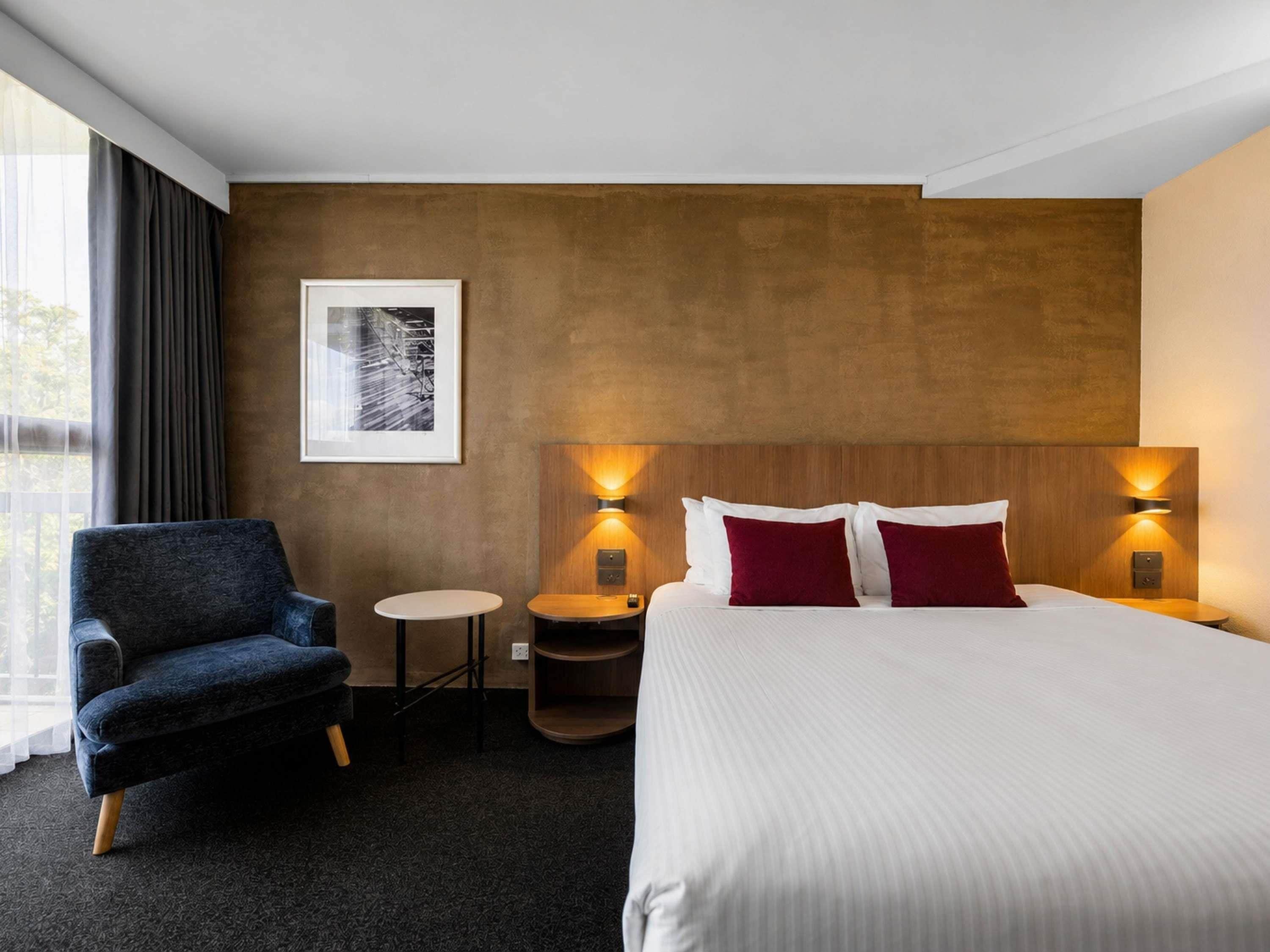 A hotel room with a bed, a chair, a small table, and a wall-mounted lamp.