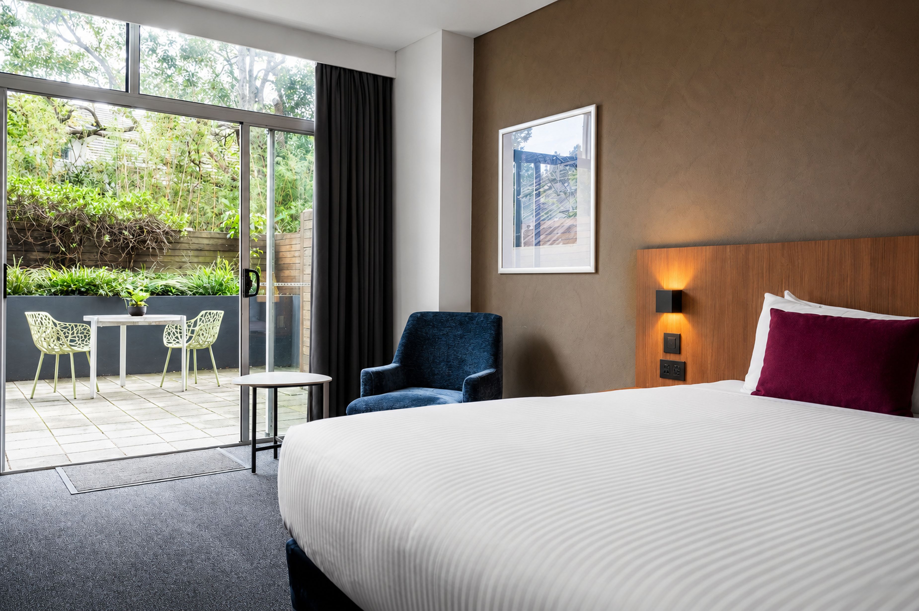 A hotel room with a large bed, a chair, a small table, and a view of greenery through the window.