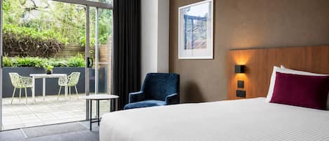 A hotel room with a large bed, a chair, a small table, and a view of greenery through the window.