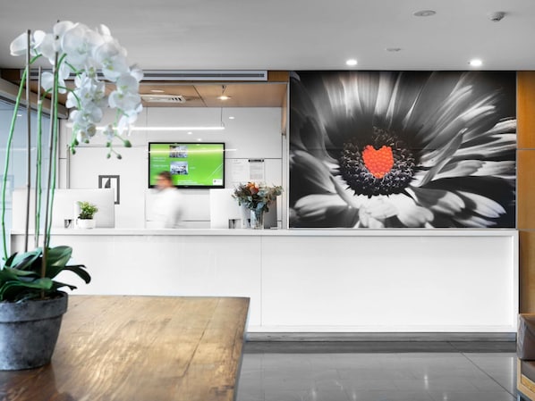 A reception desk with a digital screen, a large floral artwork, and potted plants. - Mercure Sydney St Leonards (Greenwich)