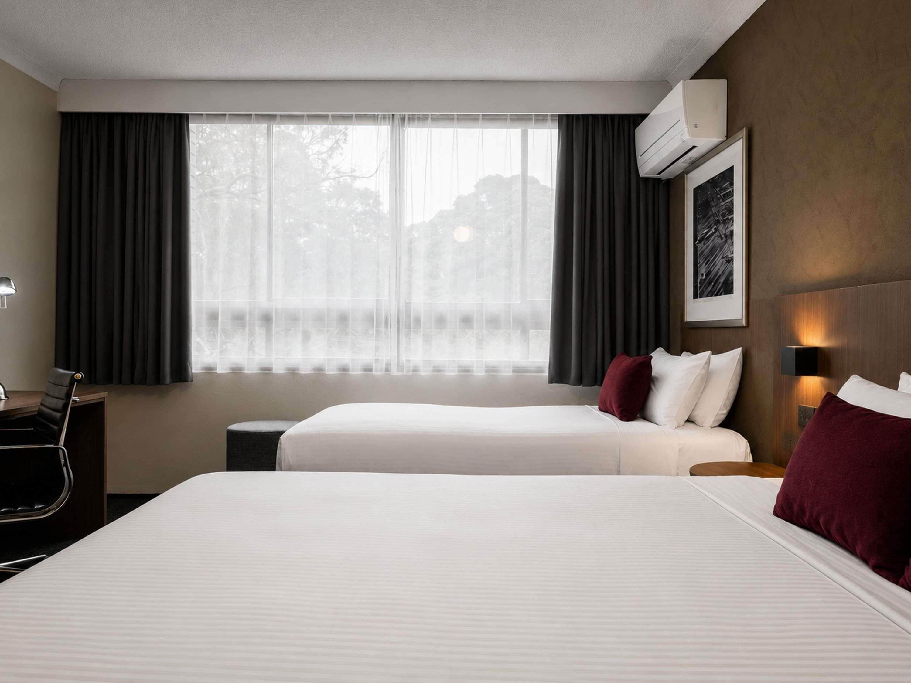 A hotel room with two beds, a desk, a chair, a window with curtains, an air conditioning unit, and a framed picture on the wall.