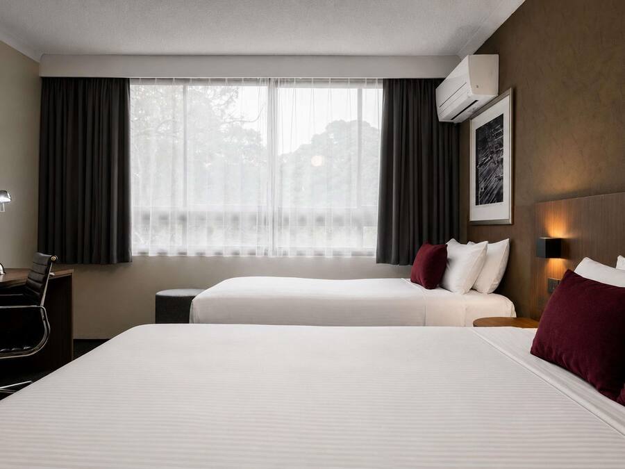 A hotel room with two beds, a desk, a chair, a window with curtains, an air conditioning unit, and a framed picture on the wall.