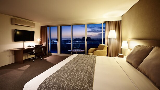 A hotel room with a large bed, a desk, a chair, a TV, a lamp, and a view of the city at dusk. - Mercure Sydney St Leonards (Greenwich)
