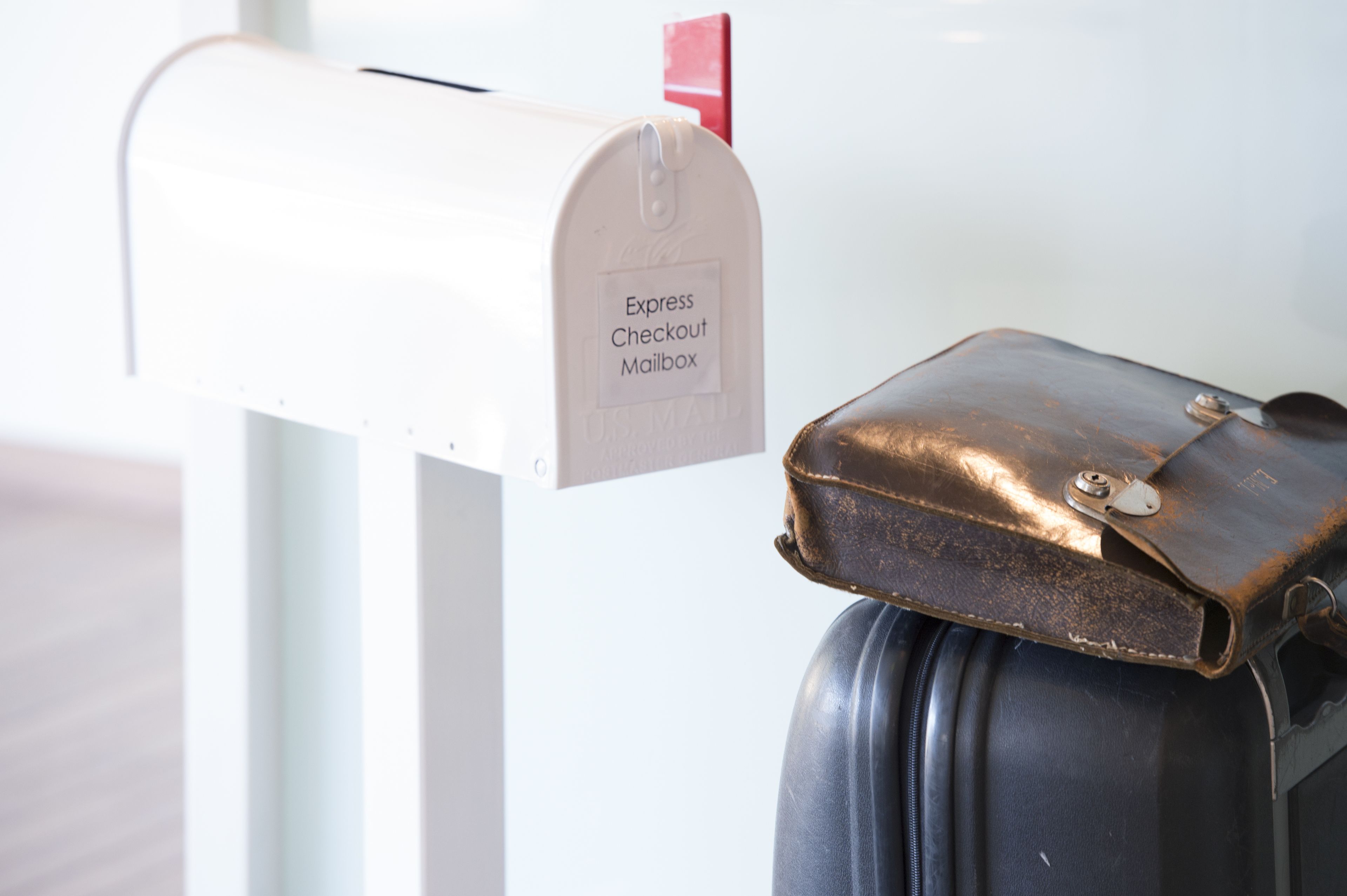 A white mailbox with an 'Express Checkout Mailbox' label, attached to a white post, next to a black suitcase.