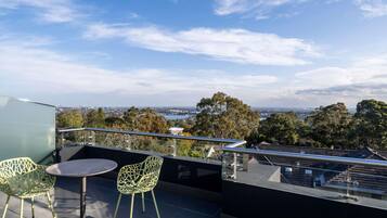 A rooftop terrace with two chairs and a table, overlooking a cityscape with trees and a bridge.