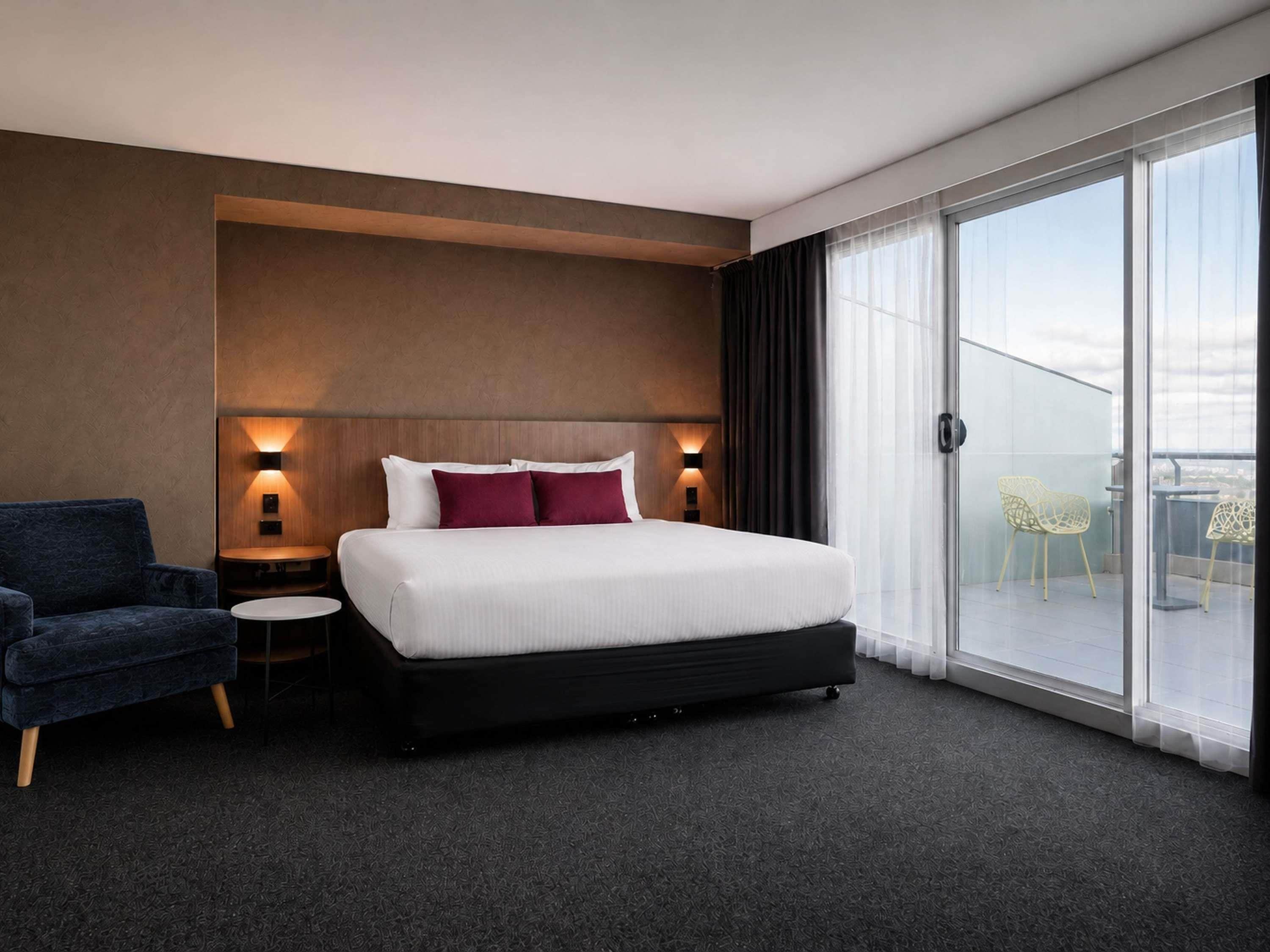 A hotel room with a large bed, a small side table, a chair, and a balcony with chairs.