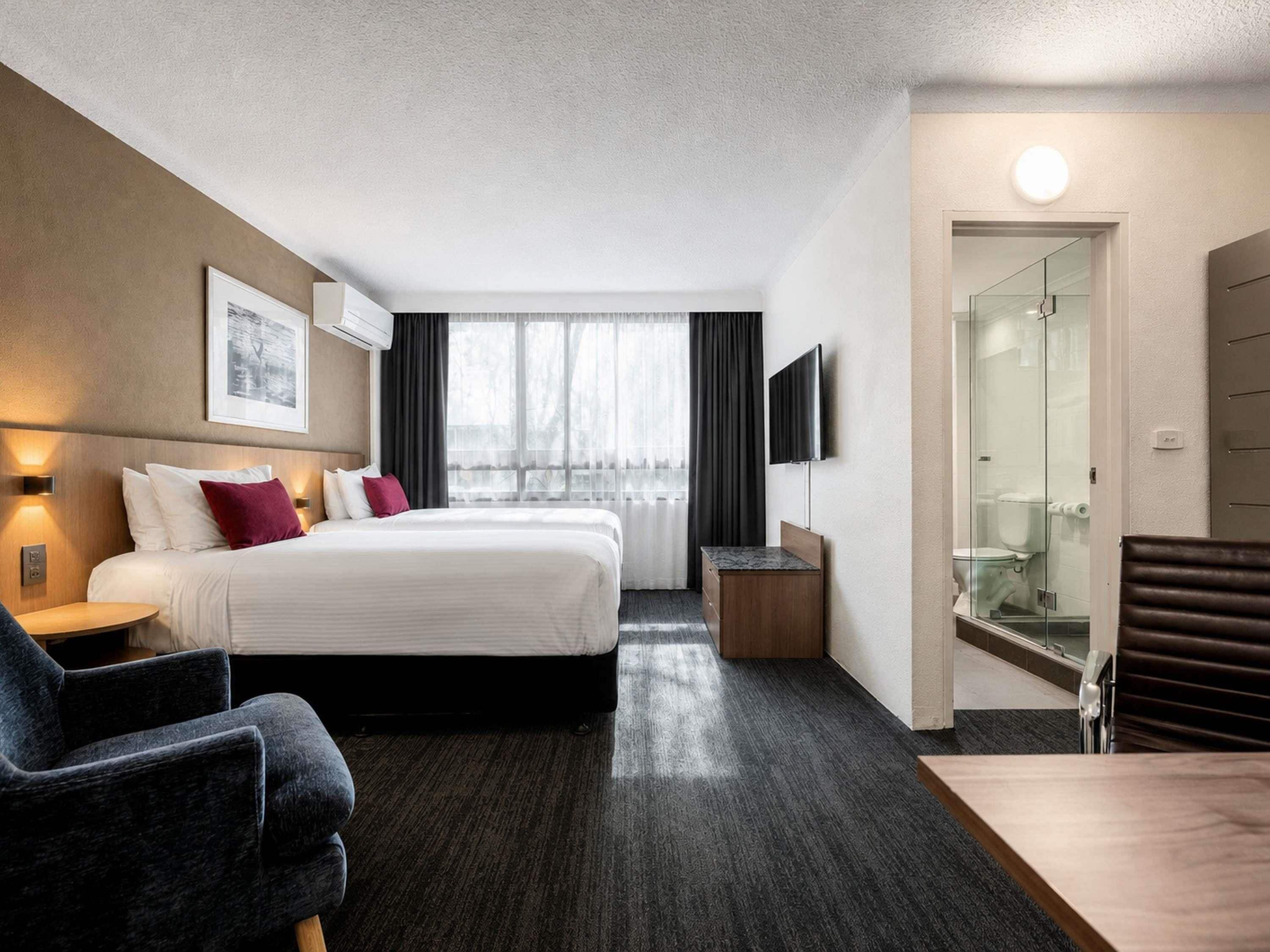 Quarto de hotel com cama, escrivaninha, cadeira, televisão e área de chuveiro com divisória de vidro.