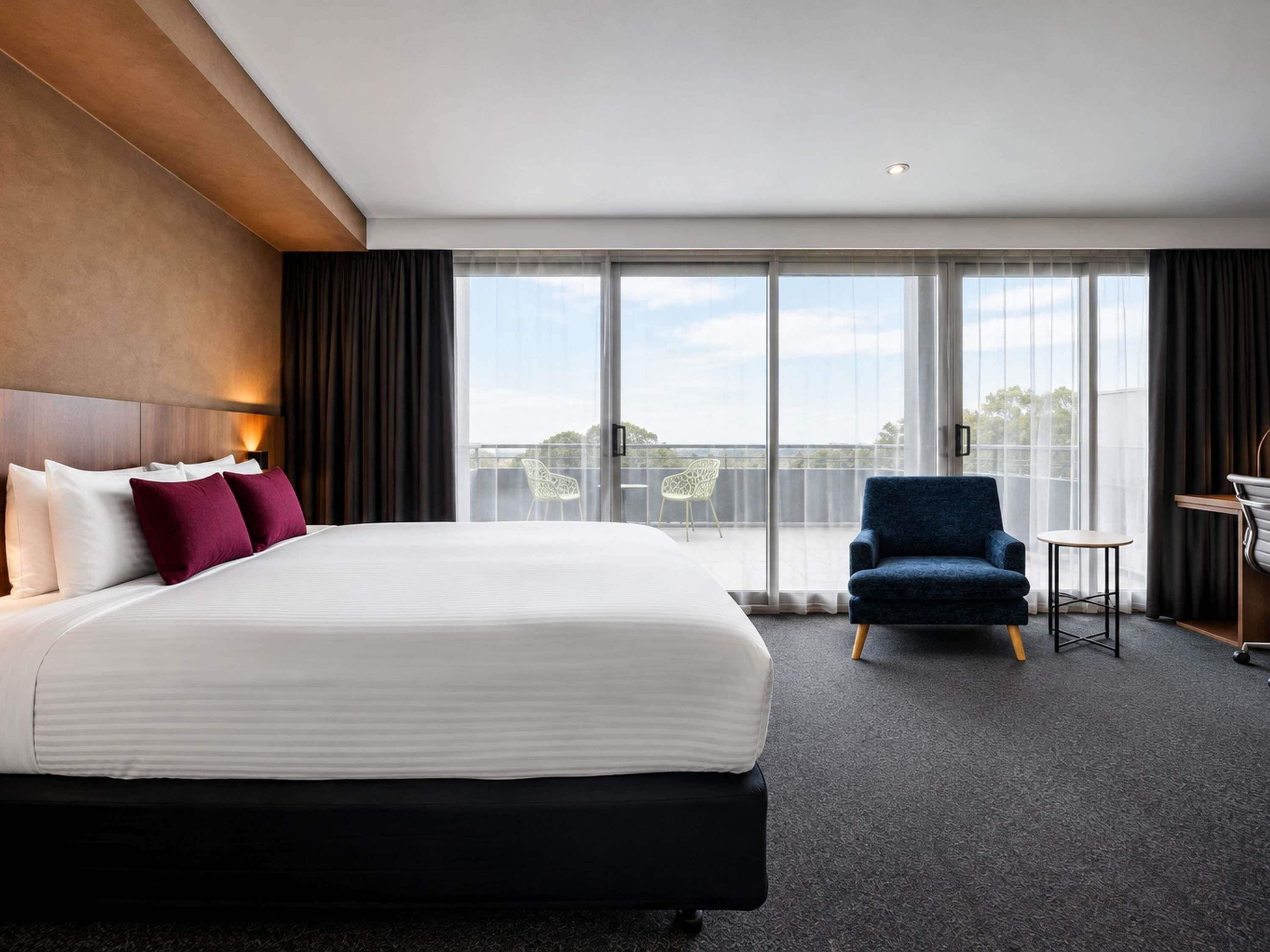 A hotel room with a large bed, a desk, a chair, and a view of the outdoors through a large window.