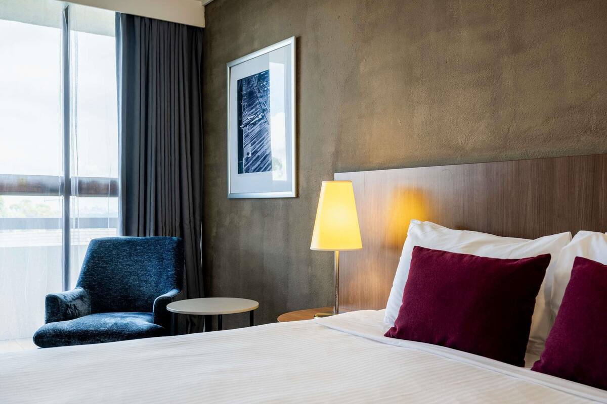 A hotel room with a bed, a chair, a small table, a lamp, and a framed picture on the wall.