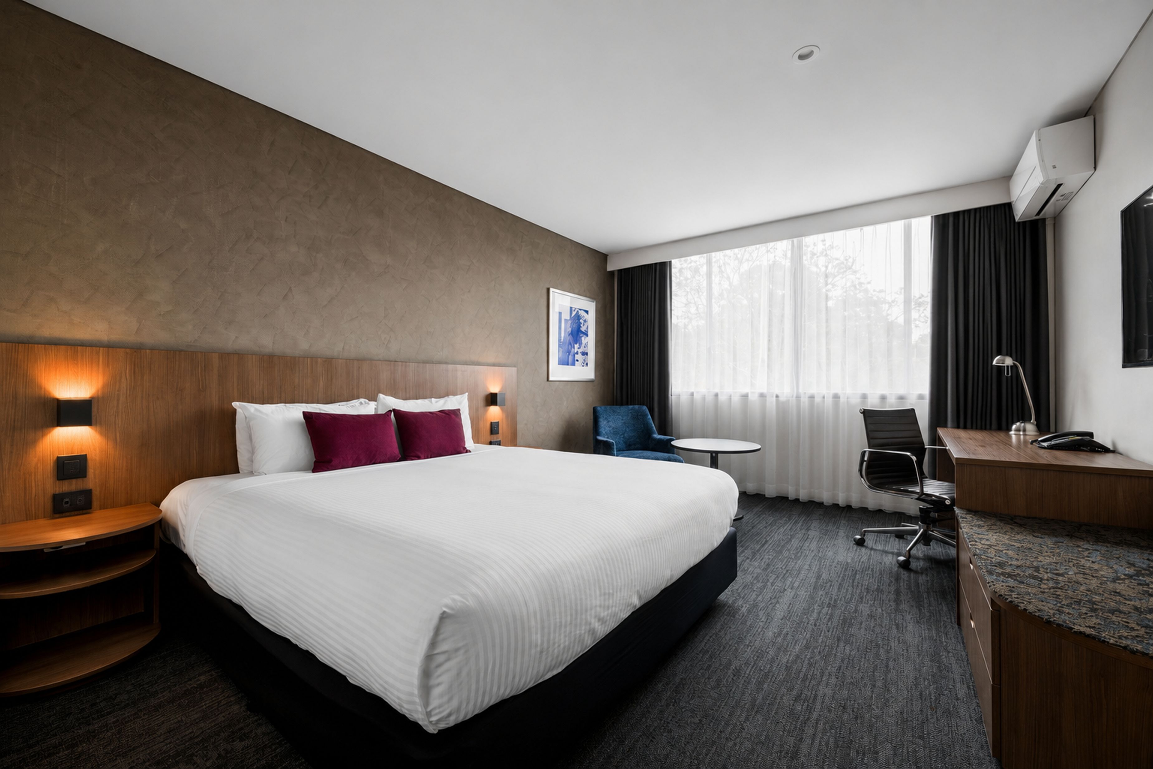A hotel room with a large bed, bedside tables, a desk, a chair, and a window with curtains.