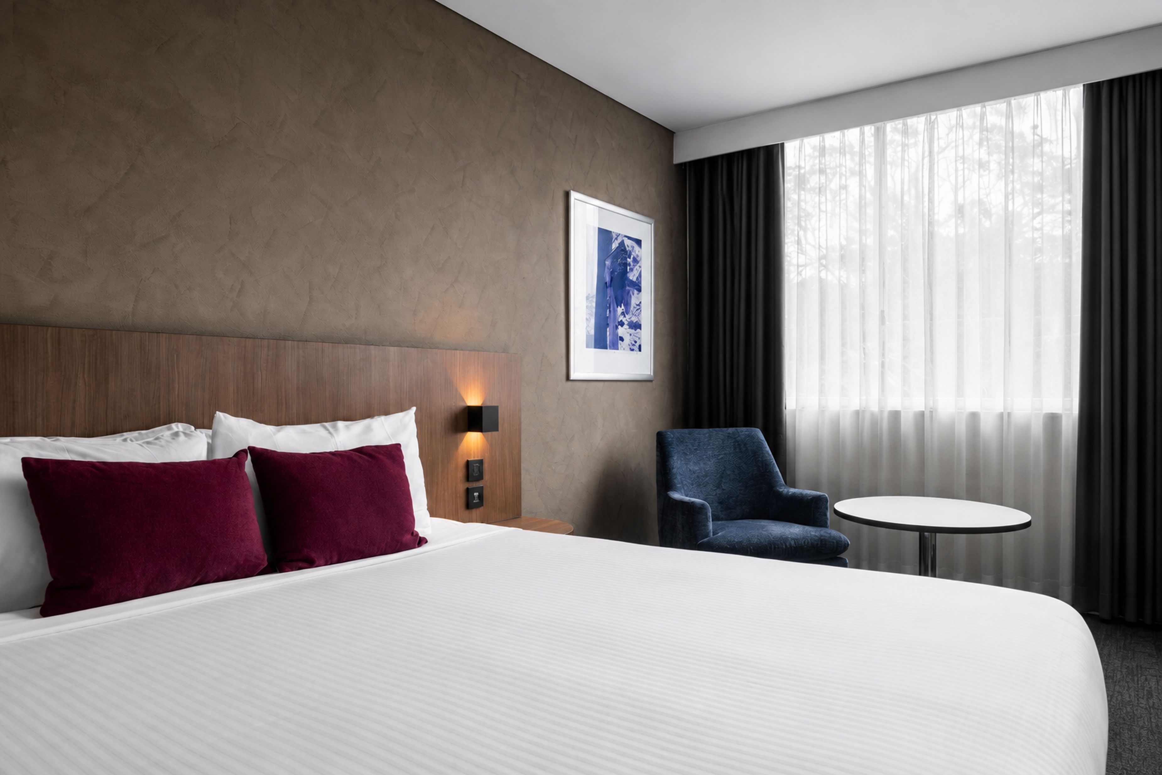 Een hotelkamer met een groot bed, een stoel, een klein tafeltje en een raam met gordijnen.