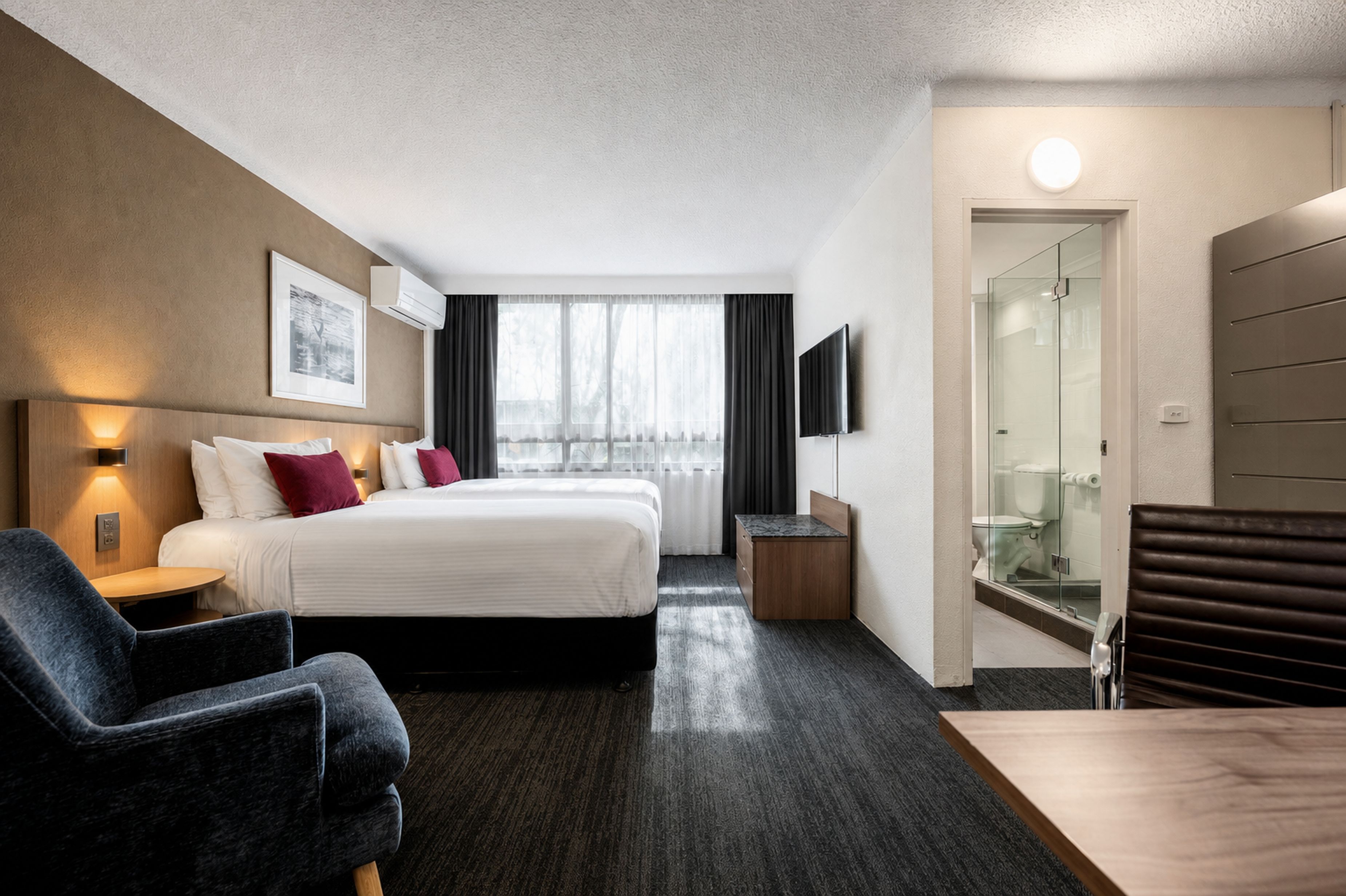 Habitación de hotel con cama, escritorio, silla, televisor y cabina de ducha de vidrio.
