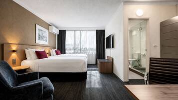 Habitación de hotel con cama, escritorio, silla, televisor y cabina de ducha de vidrio.
