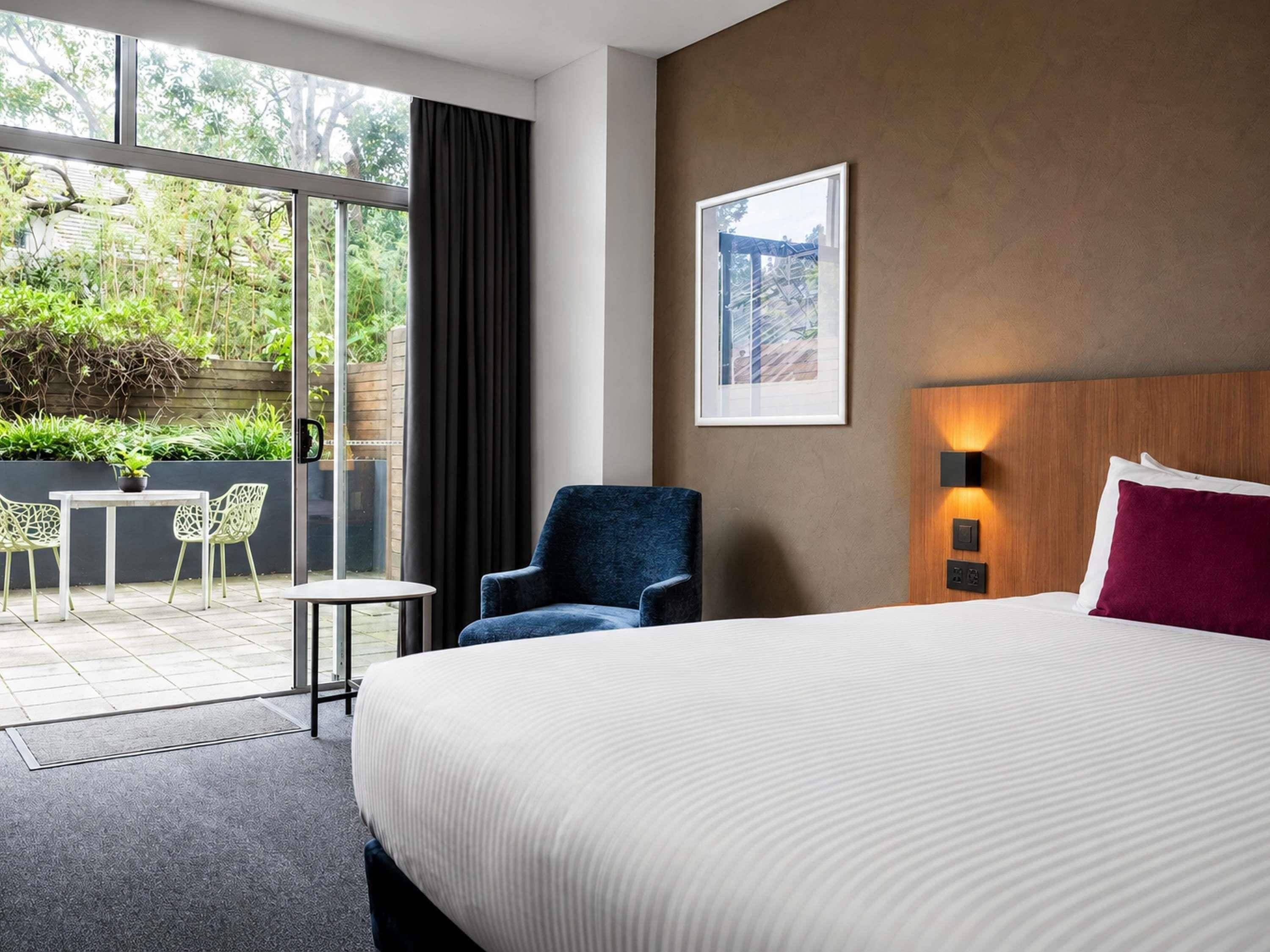 Une chambre d’hôtel avec un grand lit, une chaise, une table et une vue sur un jardin, accessible par une porte coulissante.