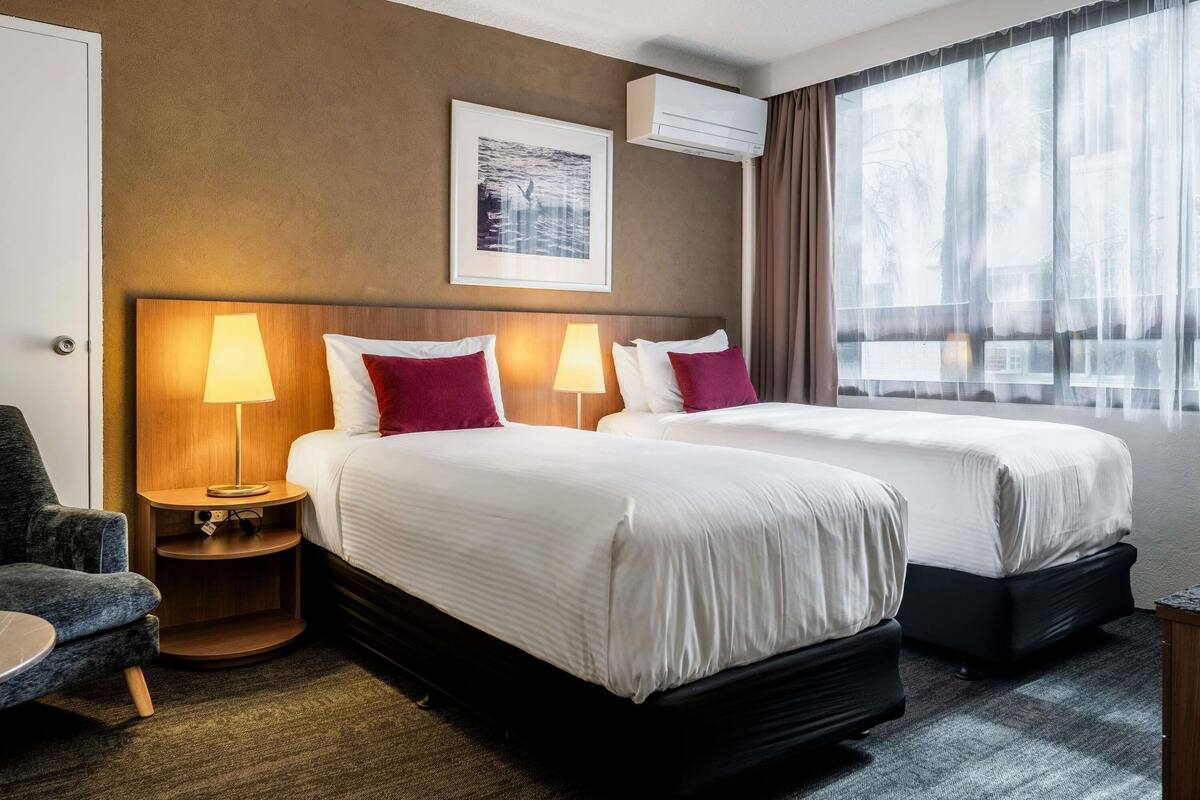 A hotel room with two beds, a headboard, a wall-mounted air conditioner, a framed picture, and a large window with curtains.