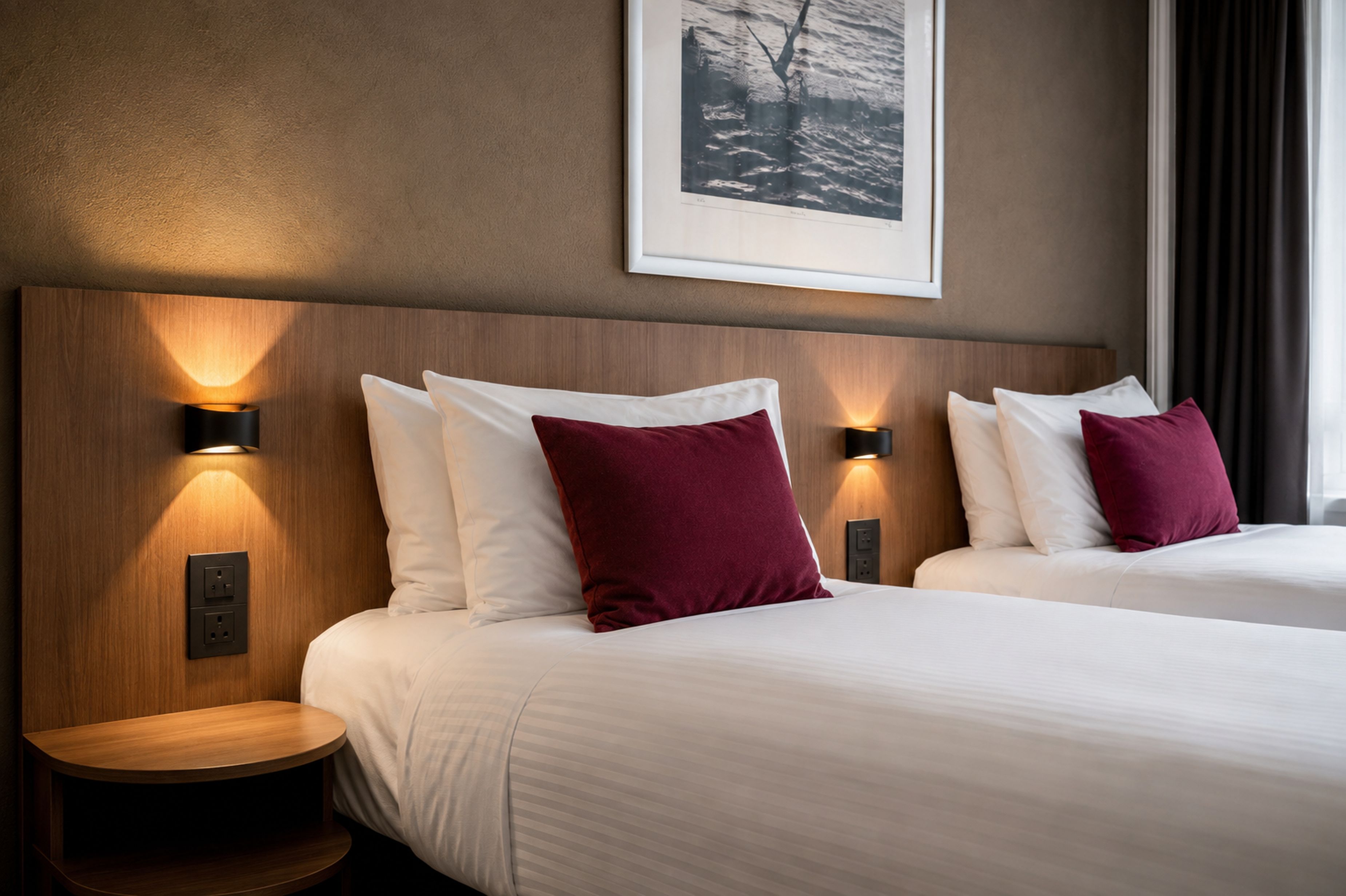 Habitación de hotel con dos camas, cada una con ropa blanca y una sola almohada roja. Hay mesitas de noche fijadas a la pared con lámparas, y un cuadro enmarcado en la pared.