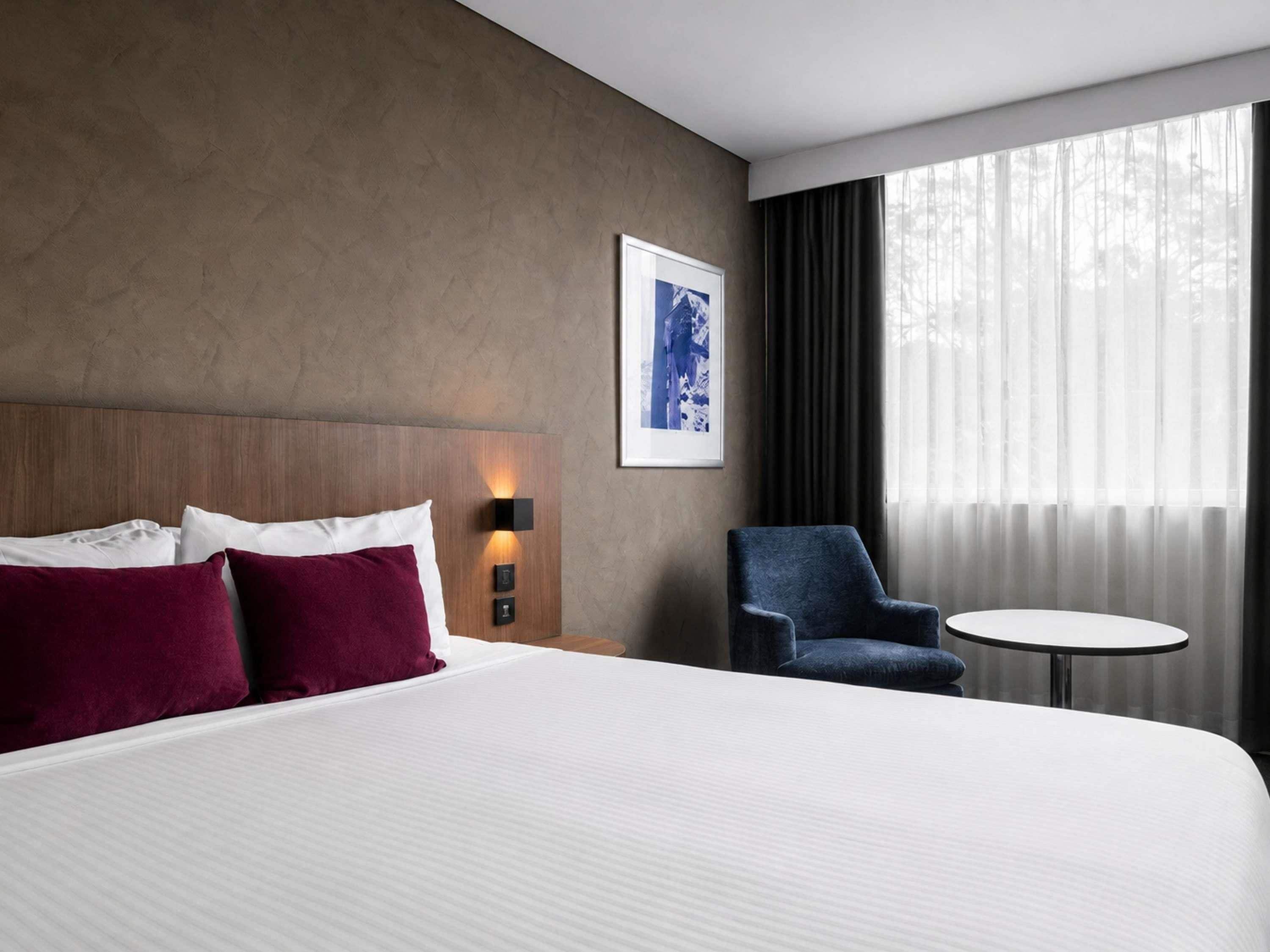 A hotel room with a large bed, a chair, a small table, and a window with curtains.