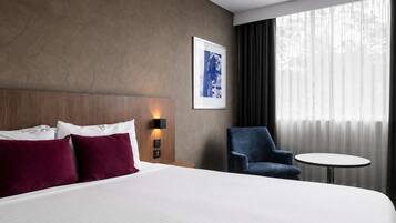 A hotel room with a large bed, a chair, a small table, and a window with curtains.
