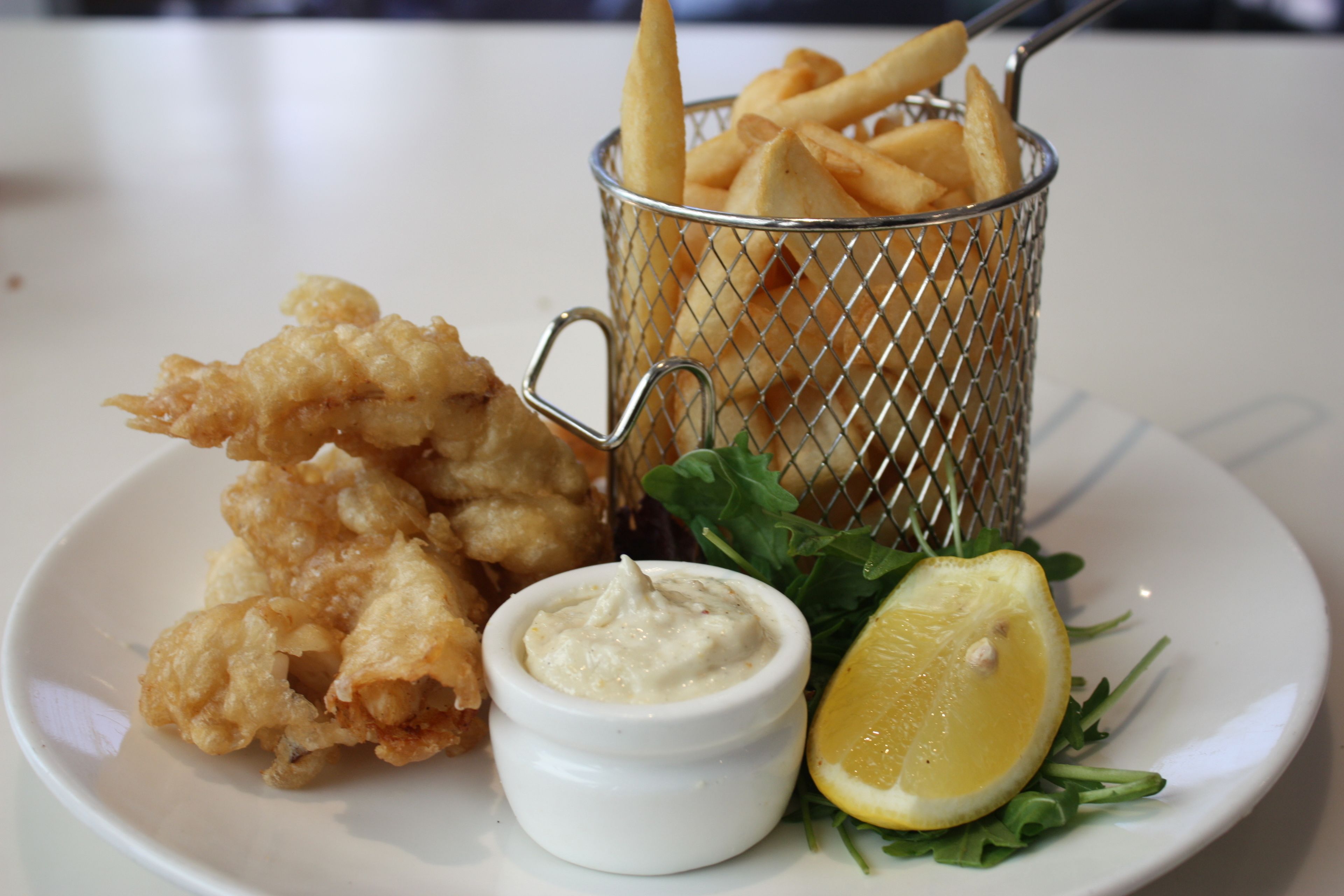 Un plato con comida frita, un recipiente pequeño con salsa para mojar, un gajo de limón y un cesto de papas fritas.