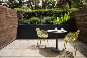 Un patio al aire libre con dos sillas amarillas y una mesa, rodeado por un muro de ladrillo y abundante vegetación.