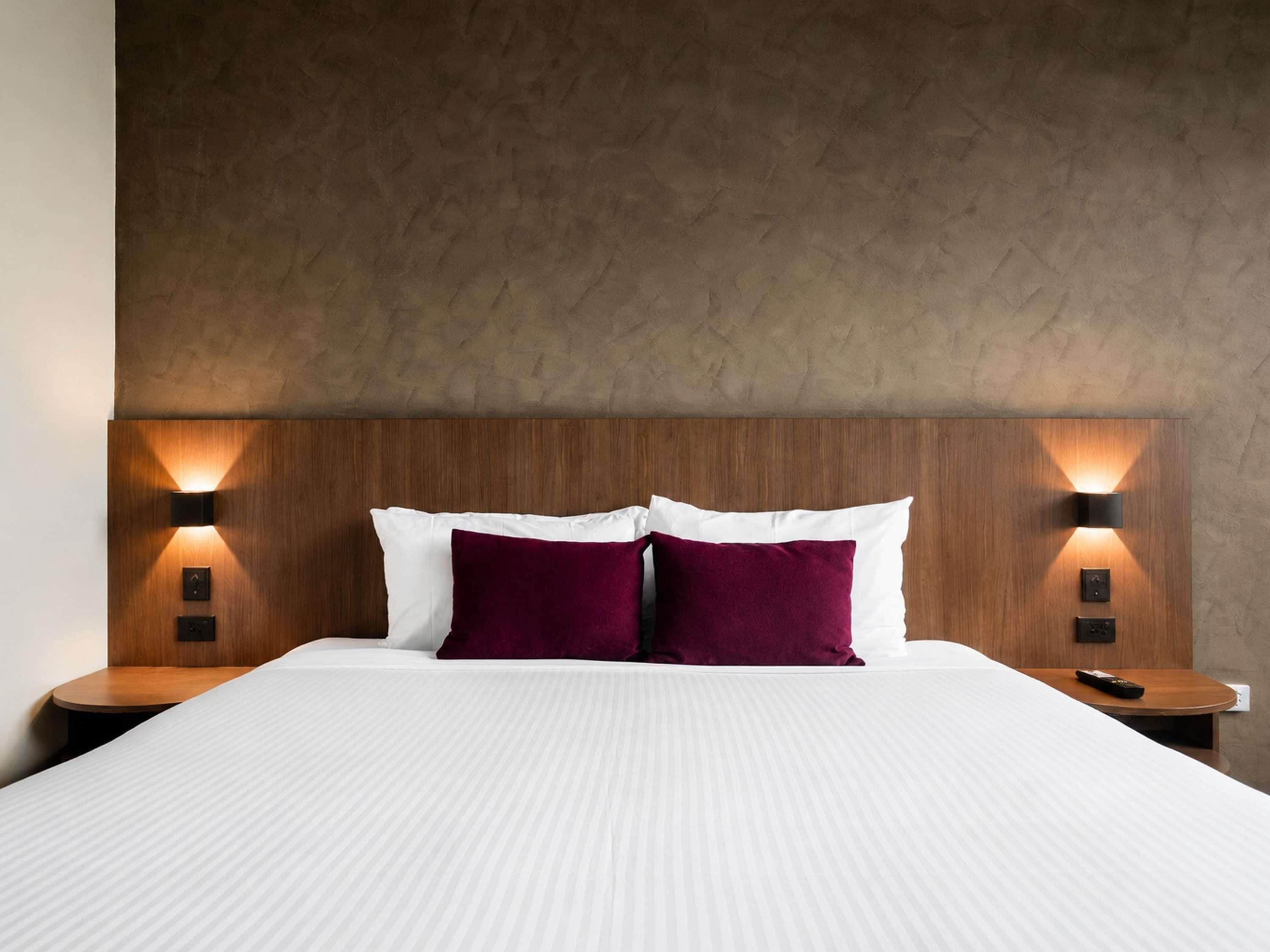 Une chambre d’hôtel avec un grand lit, deux tables de chevet, une tête de lit en bois et des lampes fixées au mur.