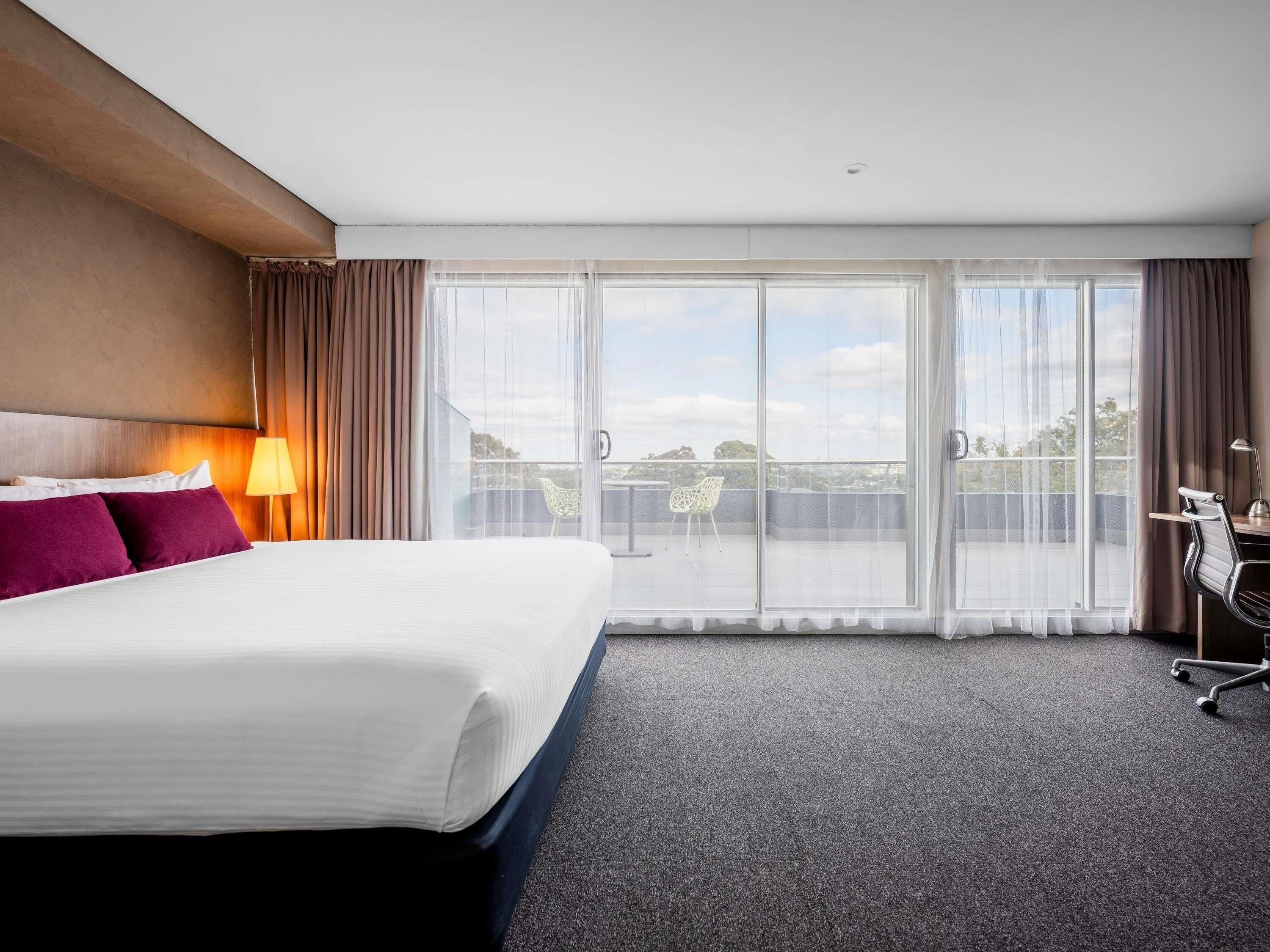 A hotel room with a bed, a desk, a chair, a lamp, and a view of the outdoors through large windows.