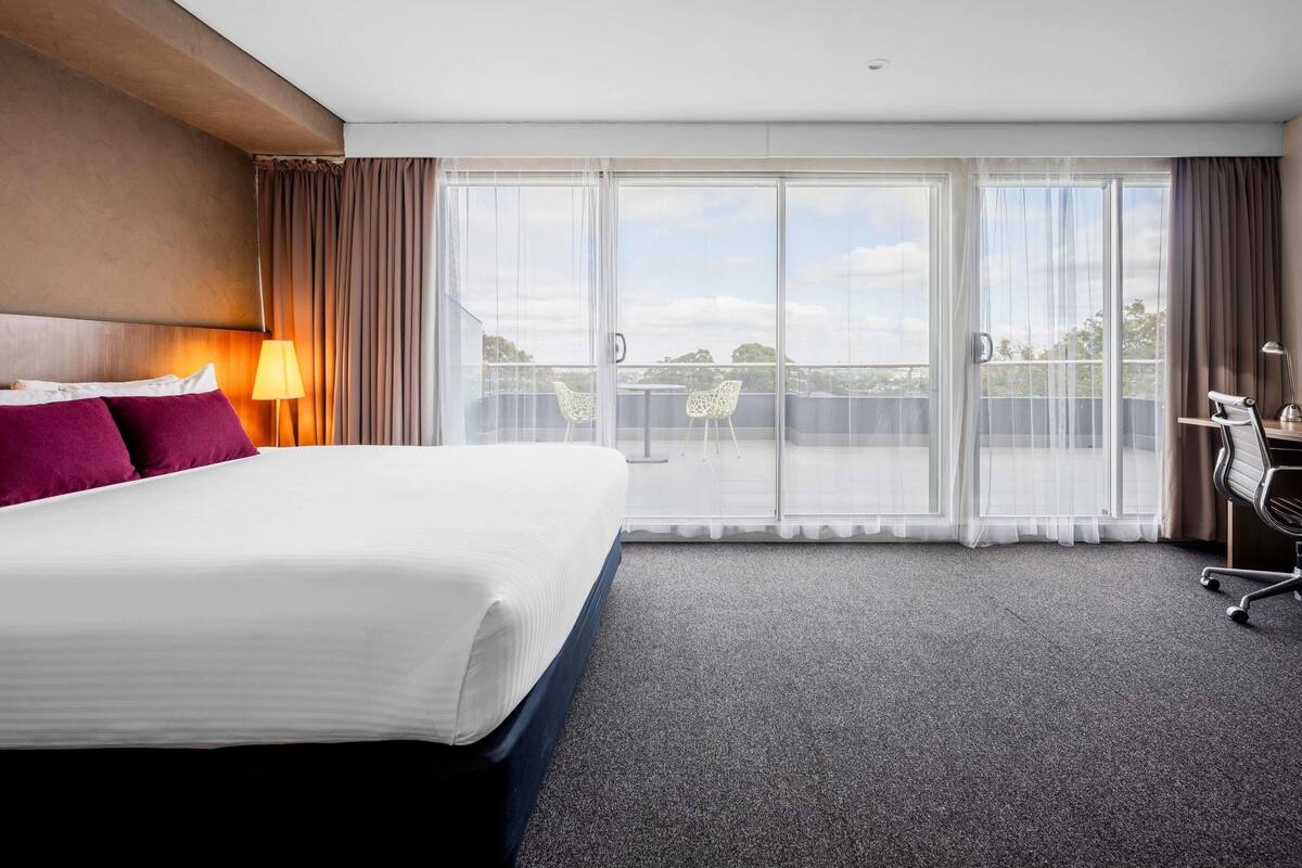 A hotel room with a bed, a desk, a chair, a lamp, and a view of the outdoors through large windows.