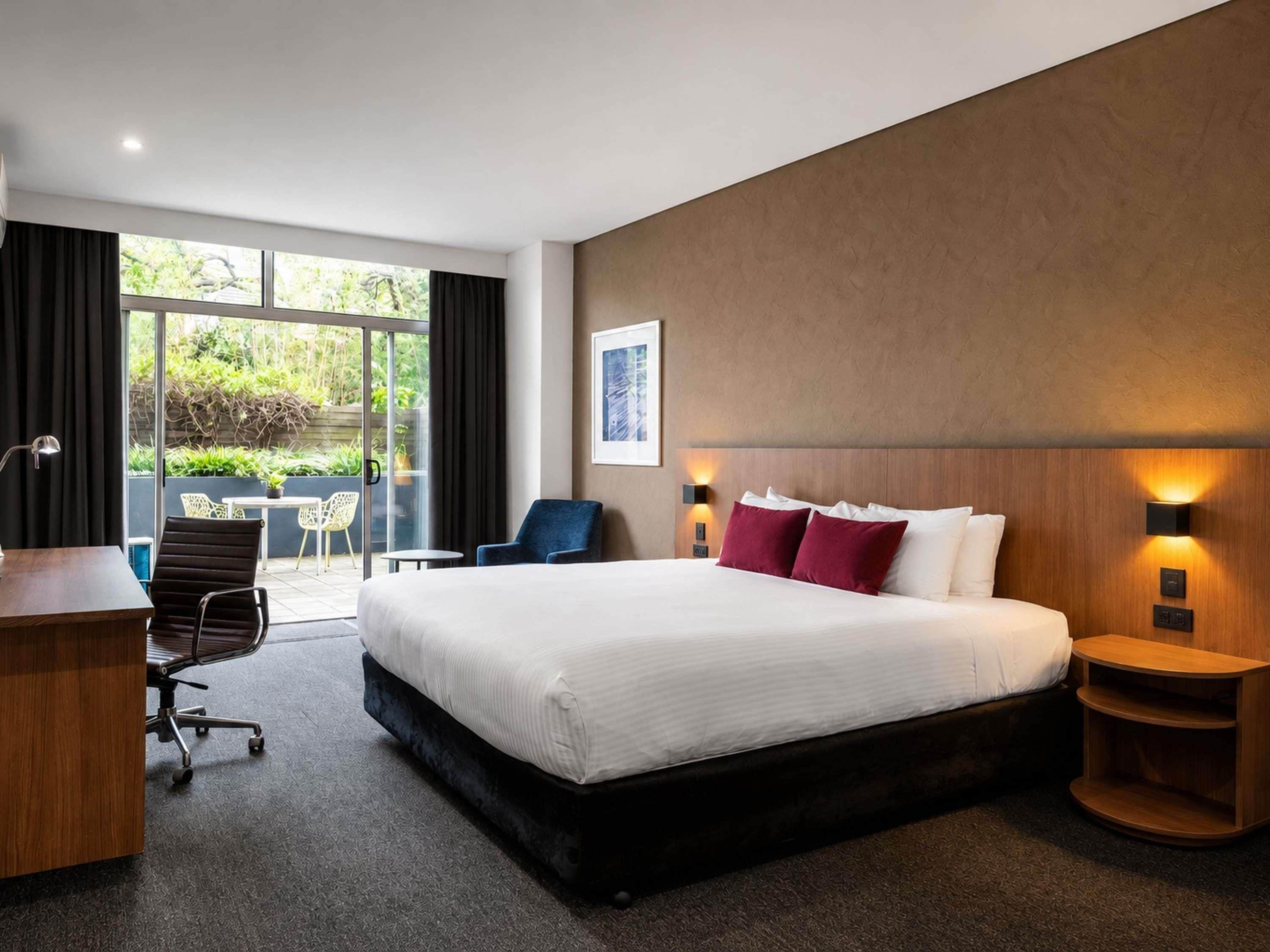 A hotel room with a large bed, a desk, a chair, a small table, and a view of the outdoors.