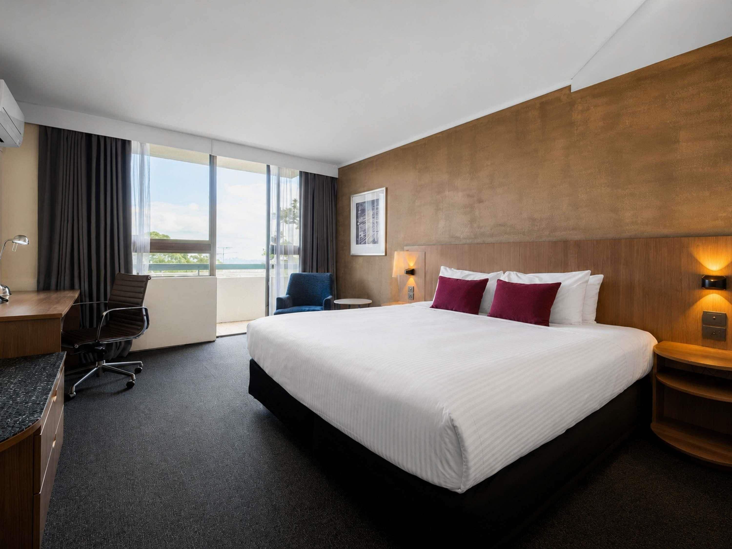 A hotel room with a large bed, a desk with a chair, a view through the window, and a wall-mounted lamp.