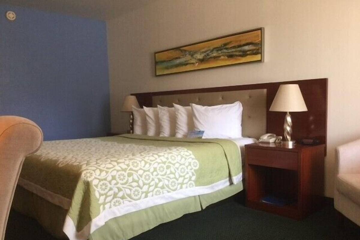 Une chambre d’hôtel avec un lit, une table de chevet, une lampe, une chaise et un tableau au mur.