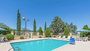 A clear blue pool with a metal ladder, surrounded by white lounge chairs and umbrellas, with trees and a fence in the background.