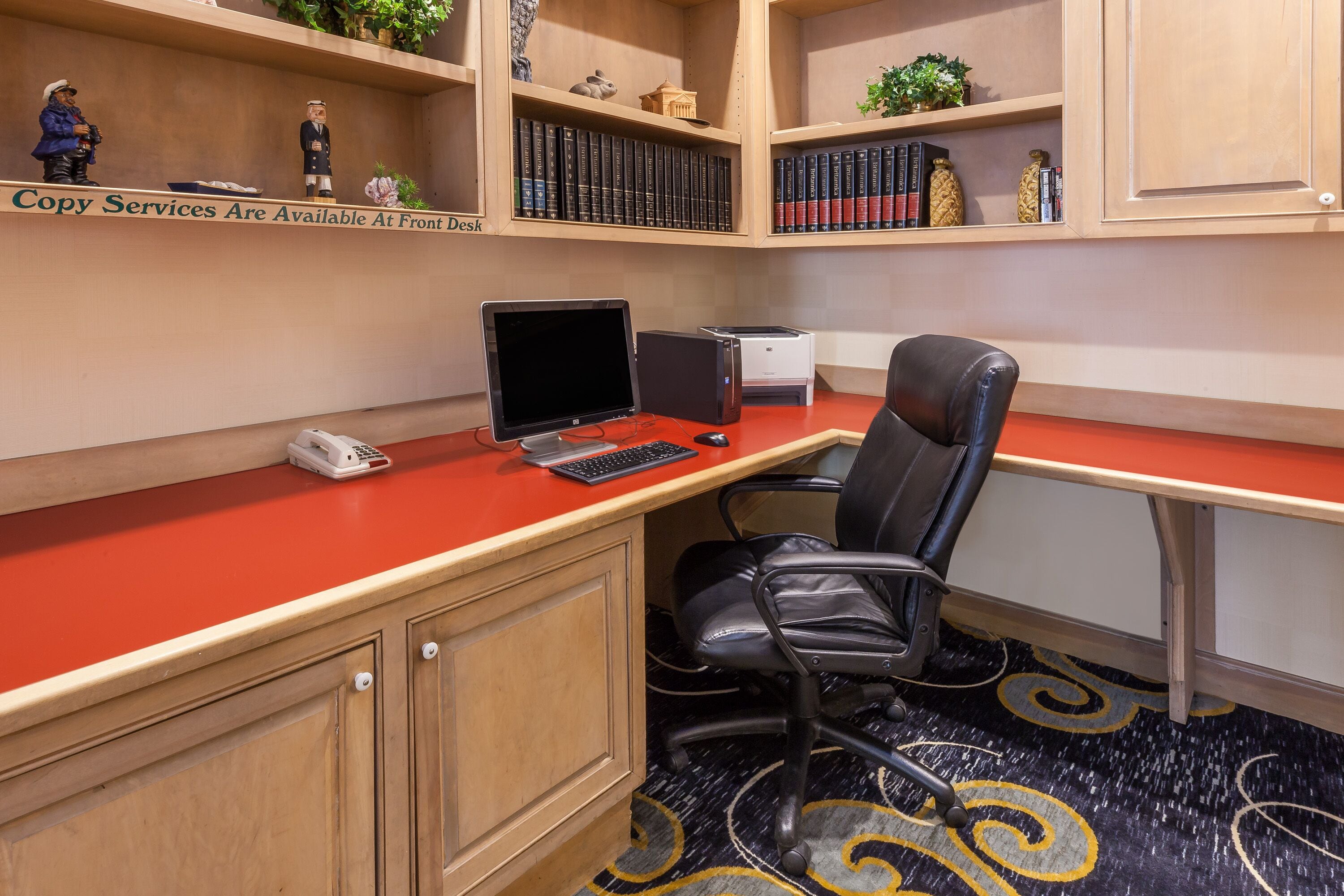 Un espace de travail comprenant un bureau, une chaise, un ordinateur, une imprimante et une bibliothèque avec des livres et des objets décoratifs.