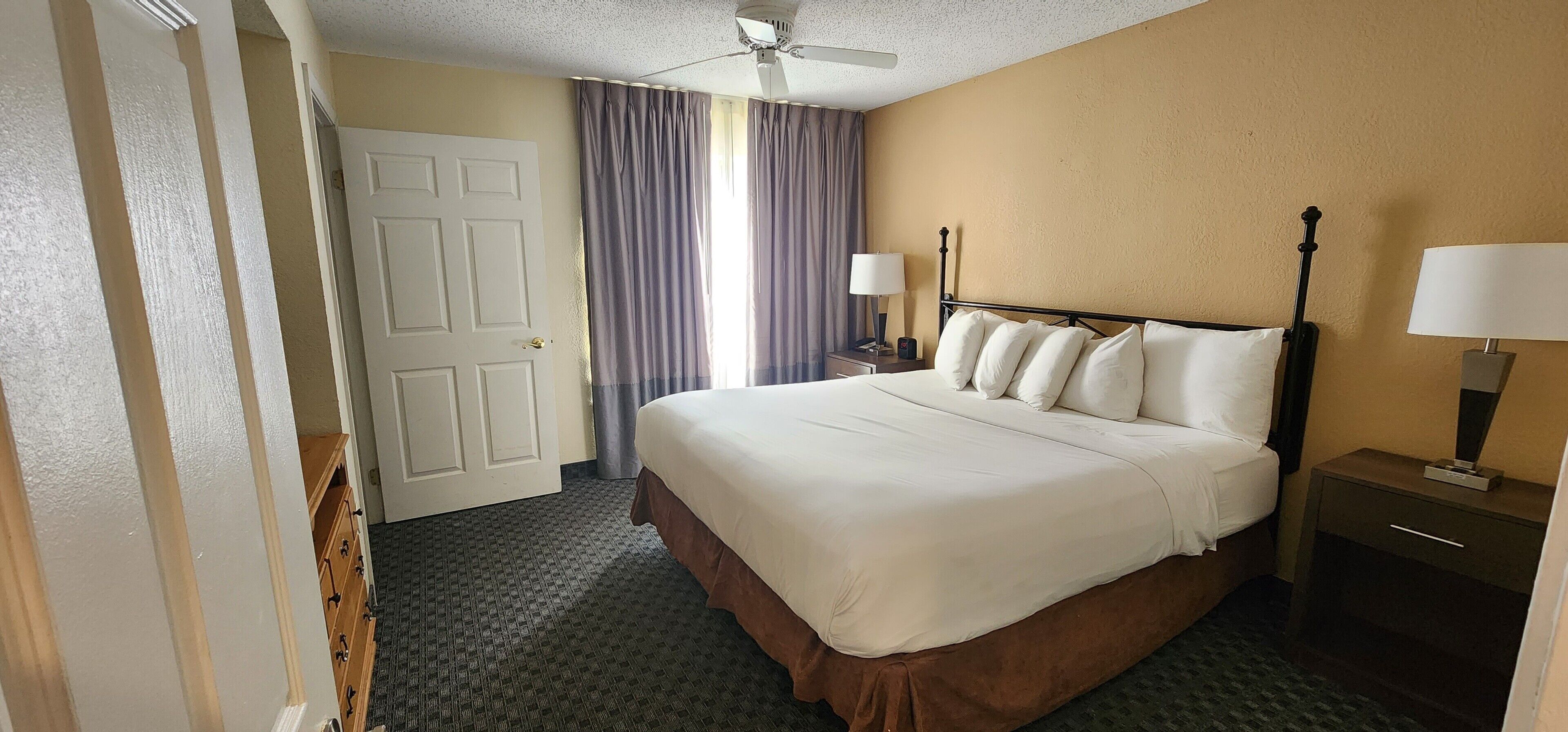 Une chambre d’hôtel avec un lit, une table de chevet avec une lampe, un ventilateur de plafond et une porte.
