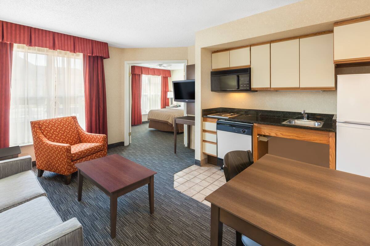 A hotel room with a kitchenette, a sofa, a chair, a small table, and a television.