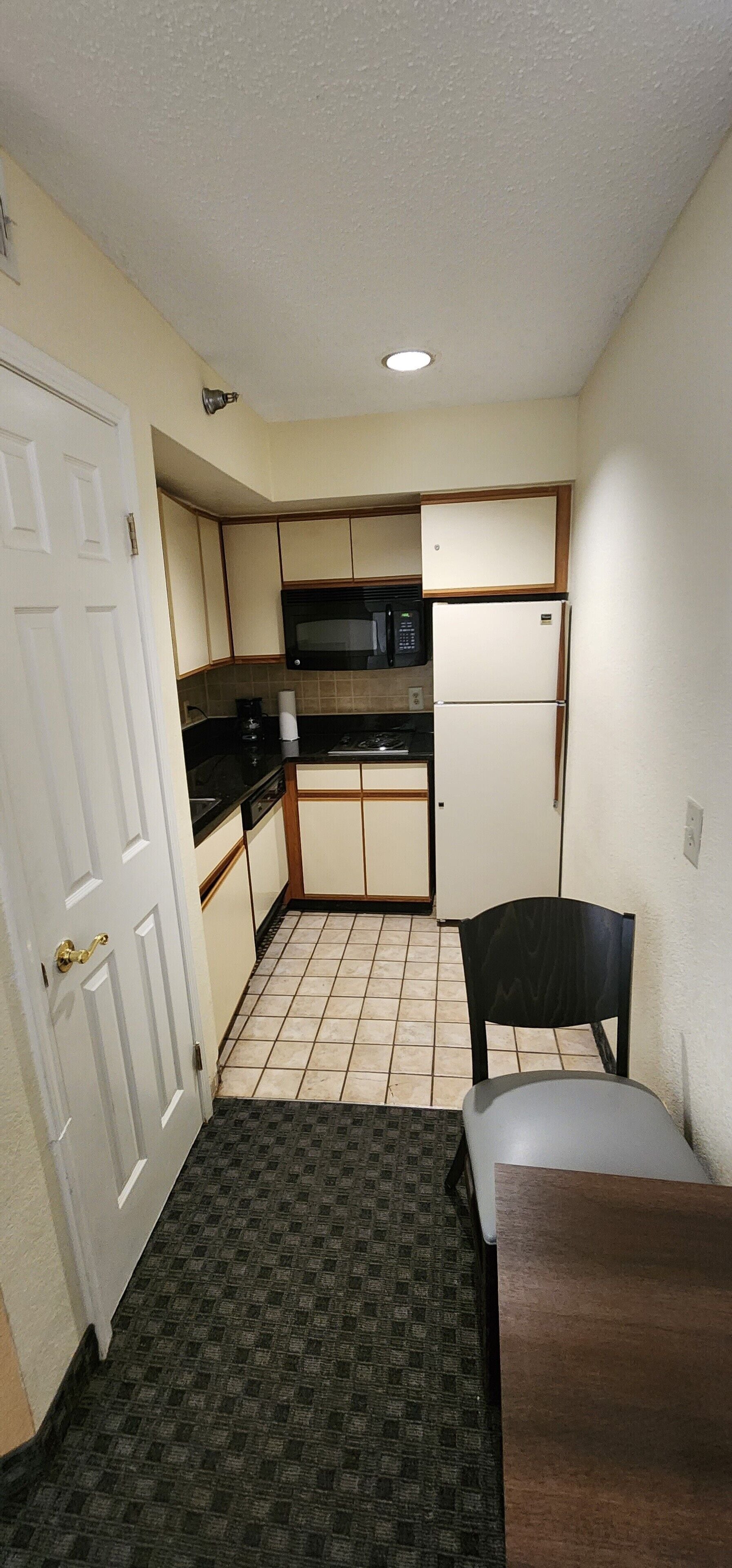 A compact kitchen with a microwave, stove, and refrigerator. There is a small dining table and a chair. The floor has a patterned carpet.