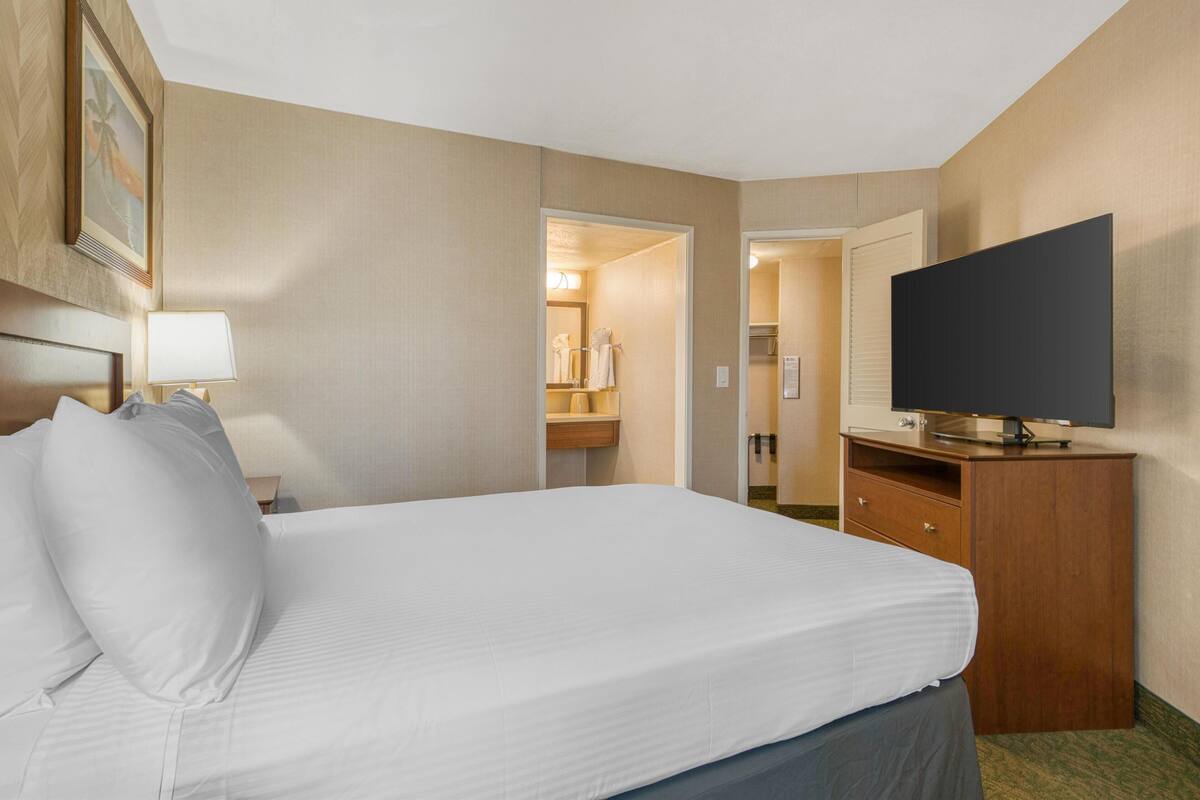 Ein Hotelzimmer mit Bett, Fernseher auf einer Holzkommode, Lampe und einem durchgehenden Türspalte sichtbaren Badezimmer.