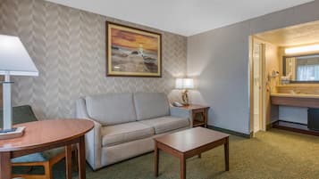 A hotel room with a sofa, a small table, a lamp, a painting of a beach scene, and a bathroom with a mirror and sink.