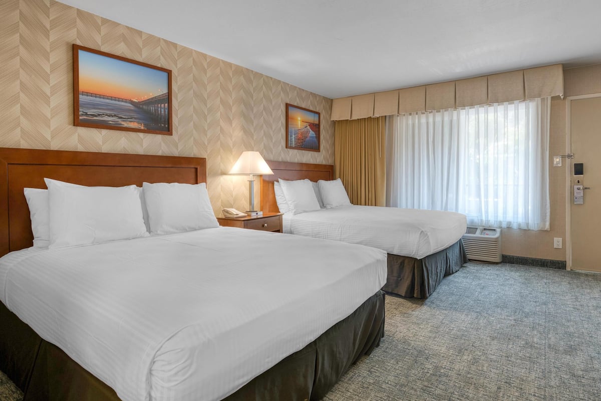 Ein Hotelzimmer mit zwei Betten, einem Nachttisch mit Lampe, einem Sonnenuntergangsbild und einem Fenster mit Vorhängen.