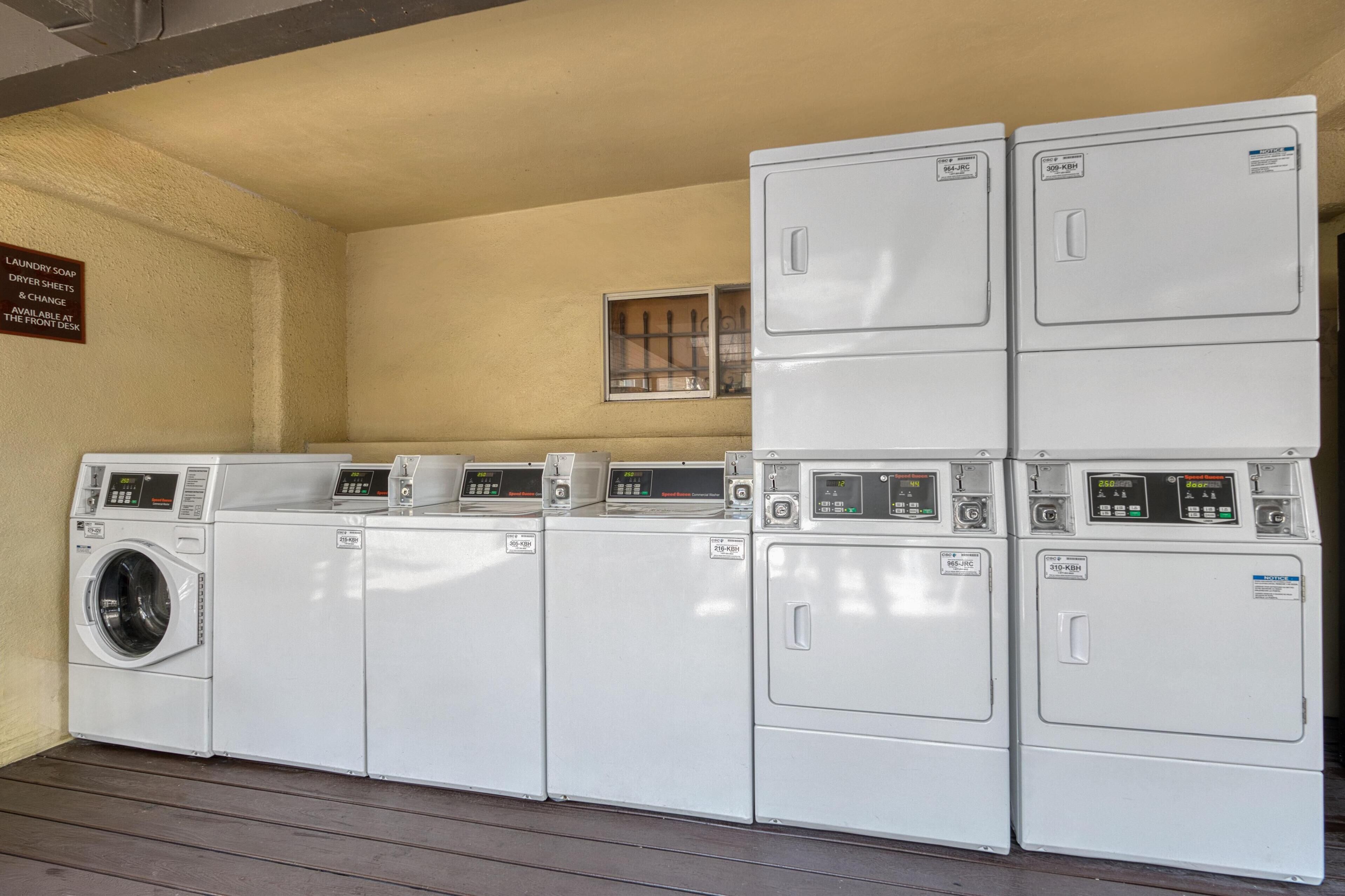 A laundry facility with multiple washing machines and dryers, a sign indicating availability of laundry supplies, and a small window above the machines.