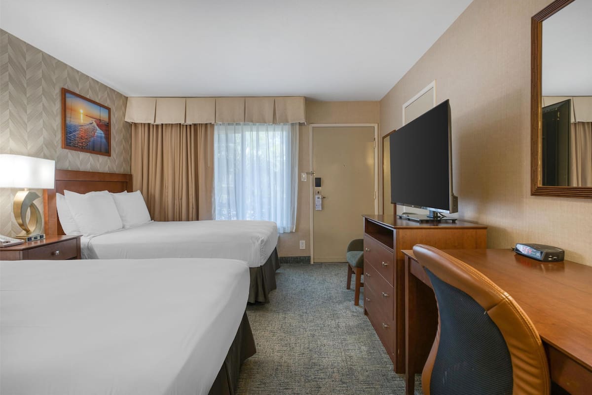 Ein Hotelzimmer mit zwei Betten, einem Schreibtisch, einem Fernseher und einem großen Fenster mit Vorhängen.